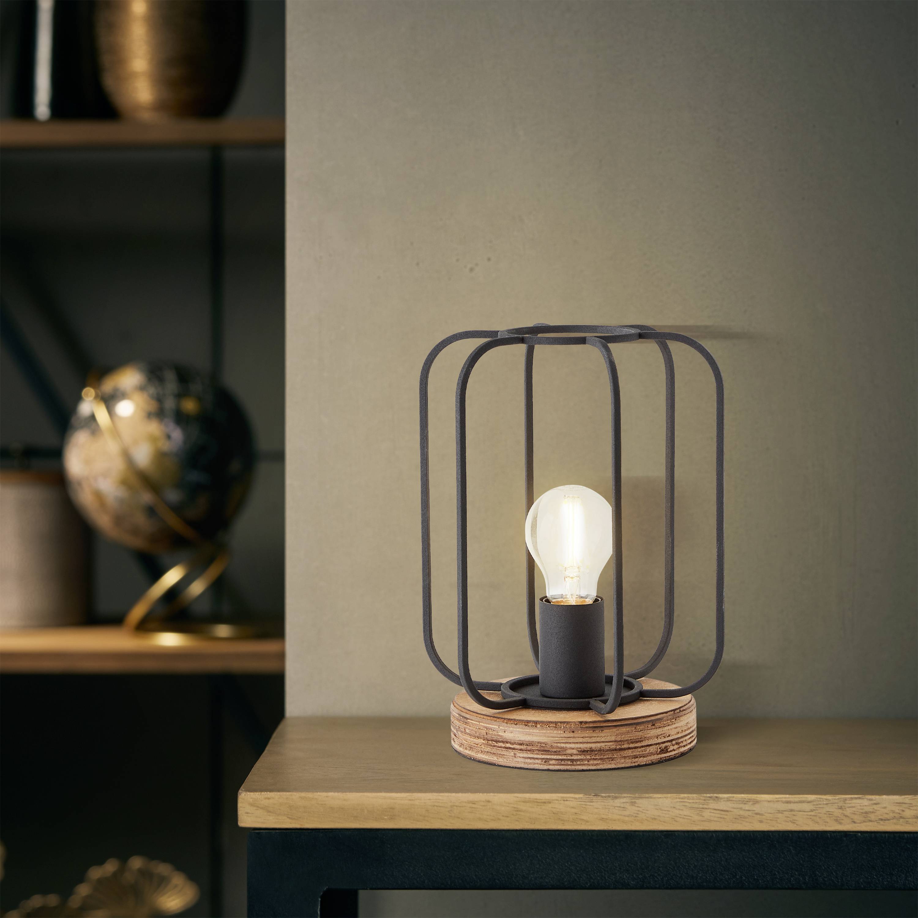 Eine Tischlampe mit Metallkäfigdesign steht auf einem Holztisch. Im Hintergrund ist ein Globus auf einem Regal sichtbar.