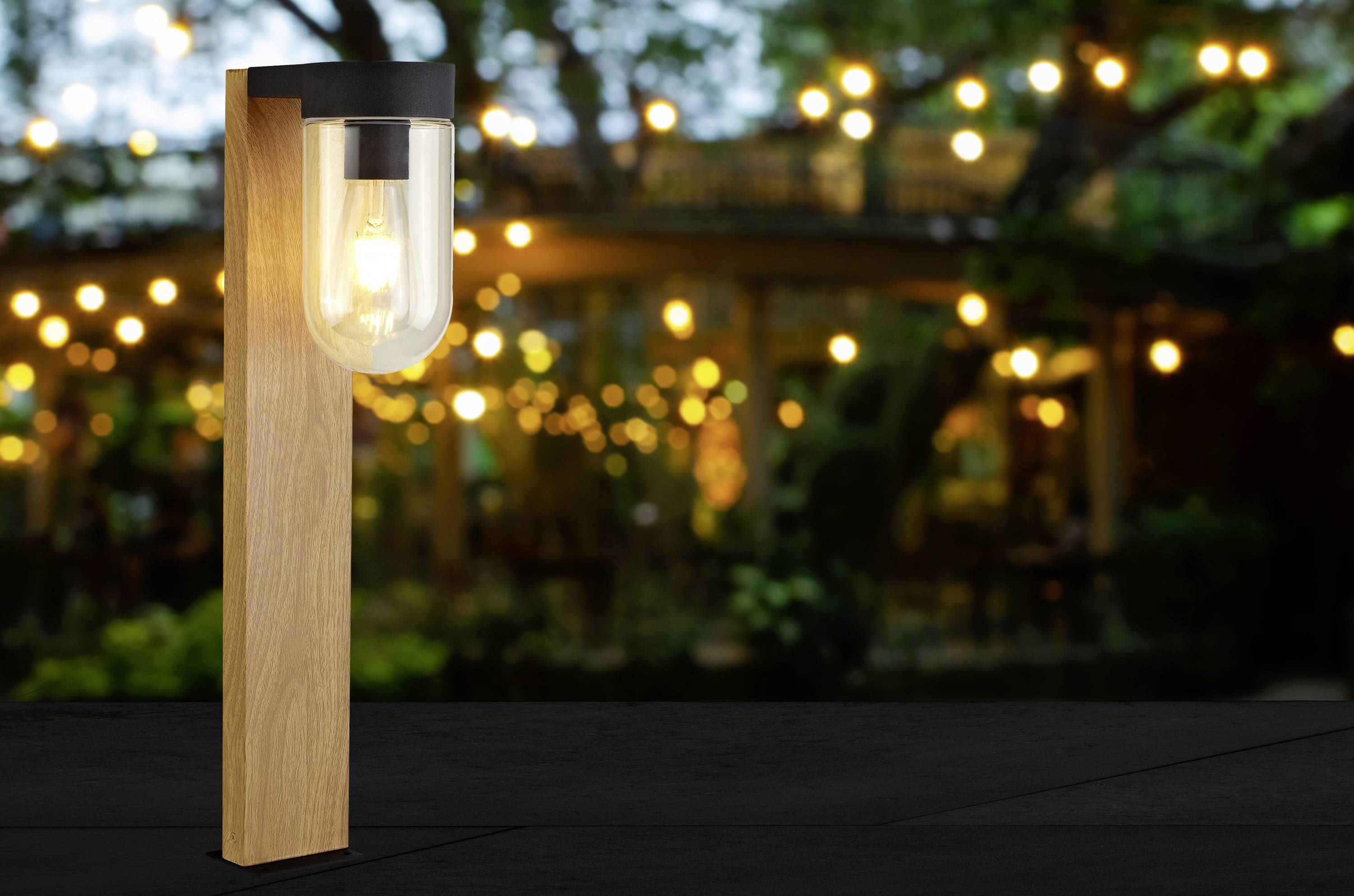 Eine moderne Lampe aus Holz und Glas leuchtet sanft, im Hintergrund verschwommene Lichter einer Gartenbeleuchtung.