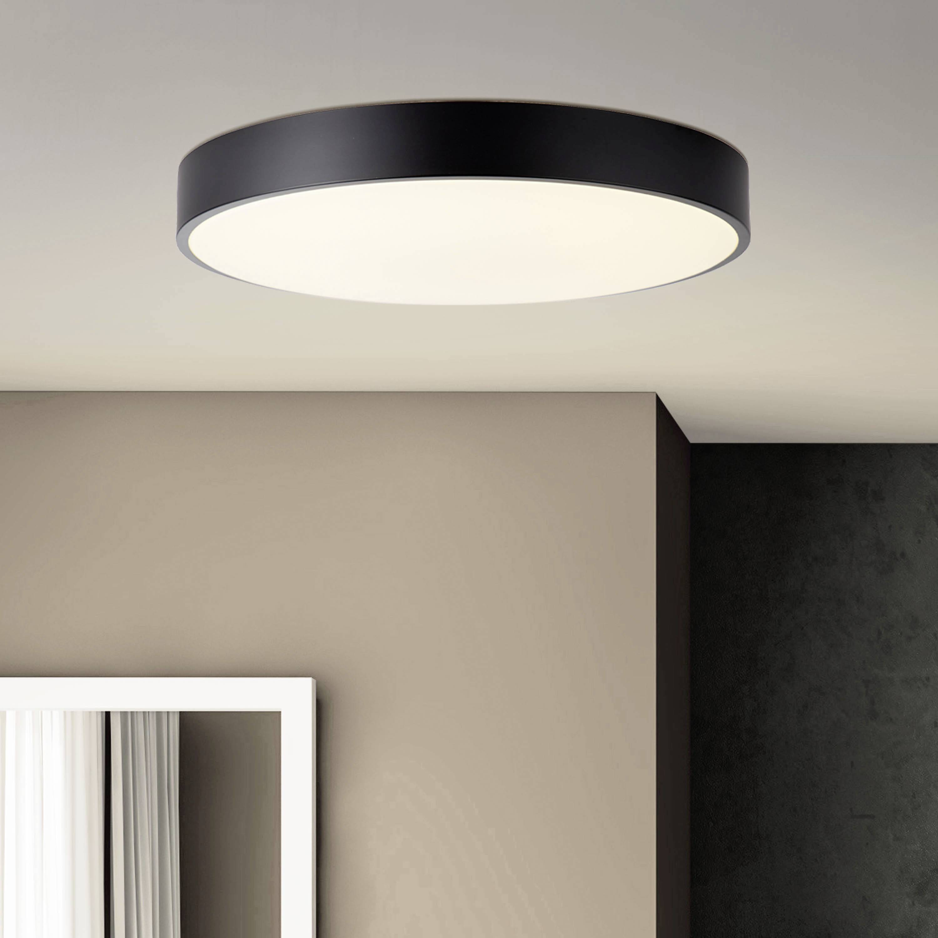 Eine moderne Deckenleuchte mit schwarzem Rand strahlt weiches Licht aus, beleuchtet einen Raum mit beiger Wand und weißem Fensterrahmen.