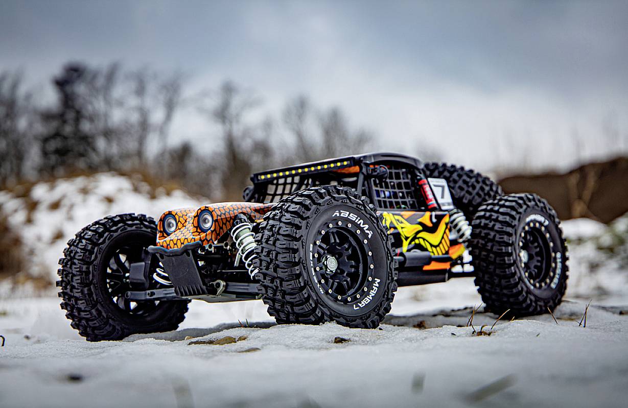 Ein ferngesteuertes Auto mit großen Reifen fährt über eine verschneite Fläche. Im Hintergrund sind Bäume schemenhaft sichtbar.