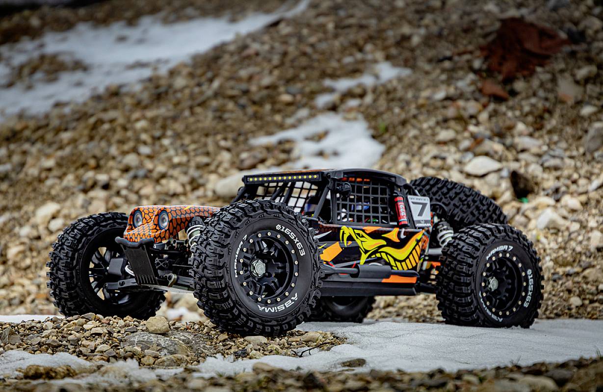 Ein ferngesteuertes Auto mit grobstolligen Reifen steht auf einem steinigen Untergrund. Es ist schwarz-orange mit einem gelben Schlangendesign.