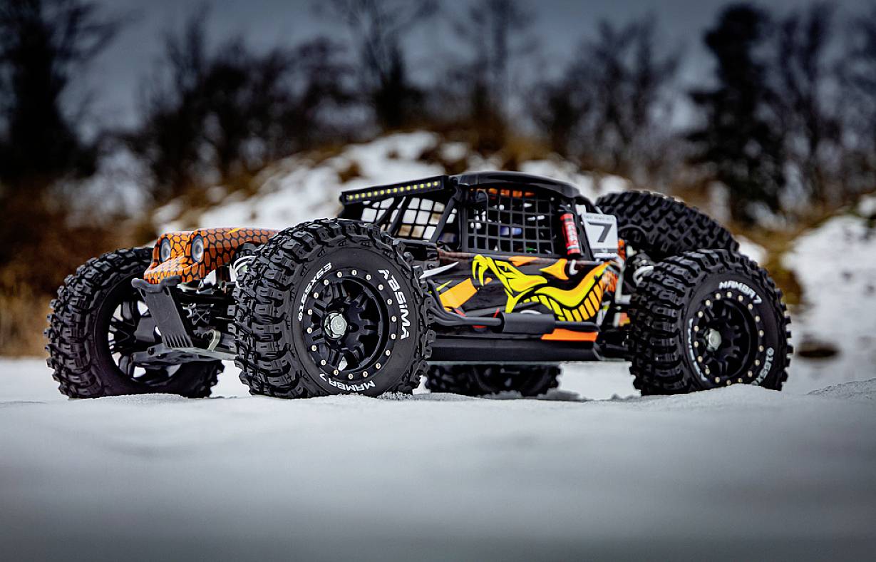 Ferngesteuertes Auto im Schnee, schwarz-orange mit auffälligem Grafikdesign an den Seiten. Grobe Reifen und schneebedeckter Hintergrund.