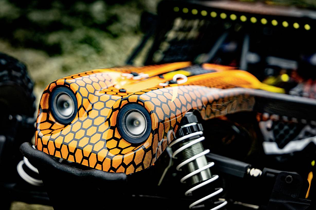 Nahaufnahme eines ferngesteuerten Geländewagens mit orange-schwarzem Wabendesign und sichtbaren Stoßdämpfern.