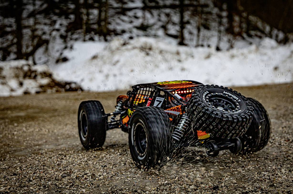 Ein ferngesteuertes Auto fährt auf einem schneebedeckten Kiesweg. Im Hintergrund sind Bäume und Schnee sichtbar.