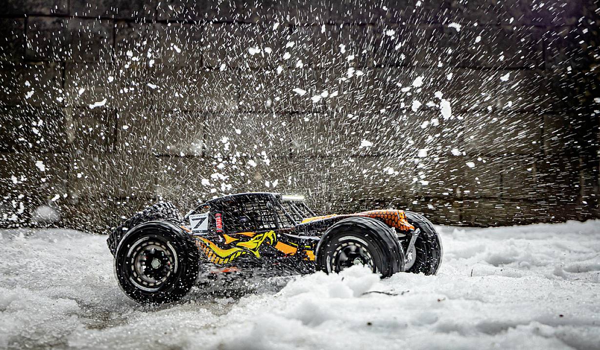 Ein ferngesteuertes Auto fährt durch Schnee, wirbelt Schneeflocken in die Luft. Der Hintergrund ist eine graue Mauer.