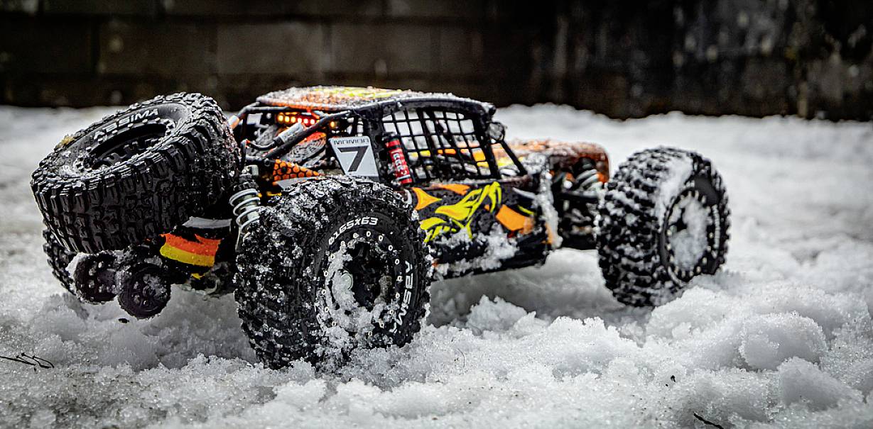 Ein ferngesteuertes Auto mit großen Reifen fährt durch Schnee, dessen Karosserie in Orange, Gelb und Schwarz gestaltet ist.