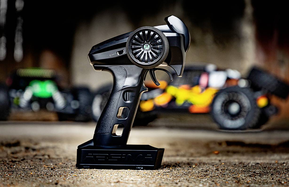 Handfernbedienung für ein RC-Auto im Vordergrund, im Hintergrund zwei unscharfe ferngesteuerte Autos auf einem rauen Untergrund.