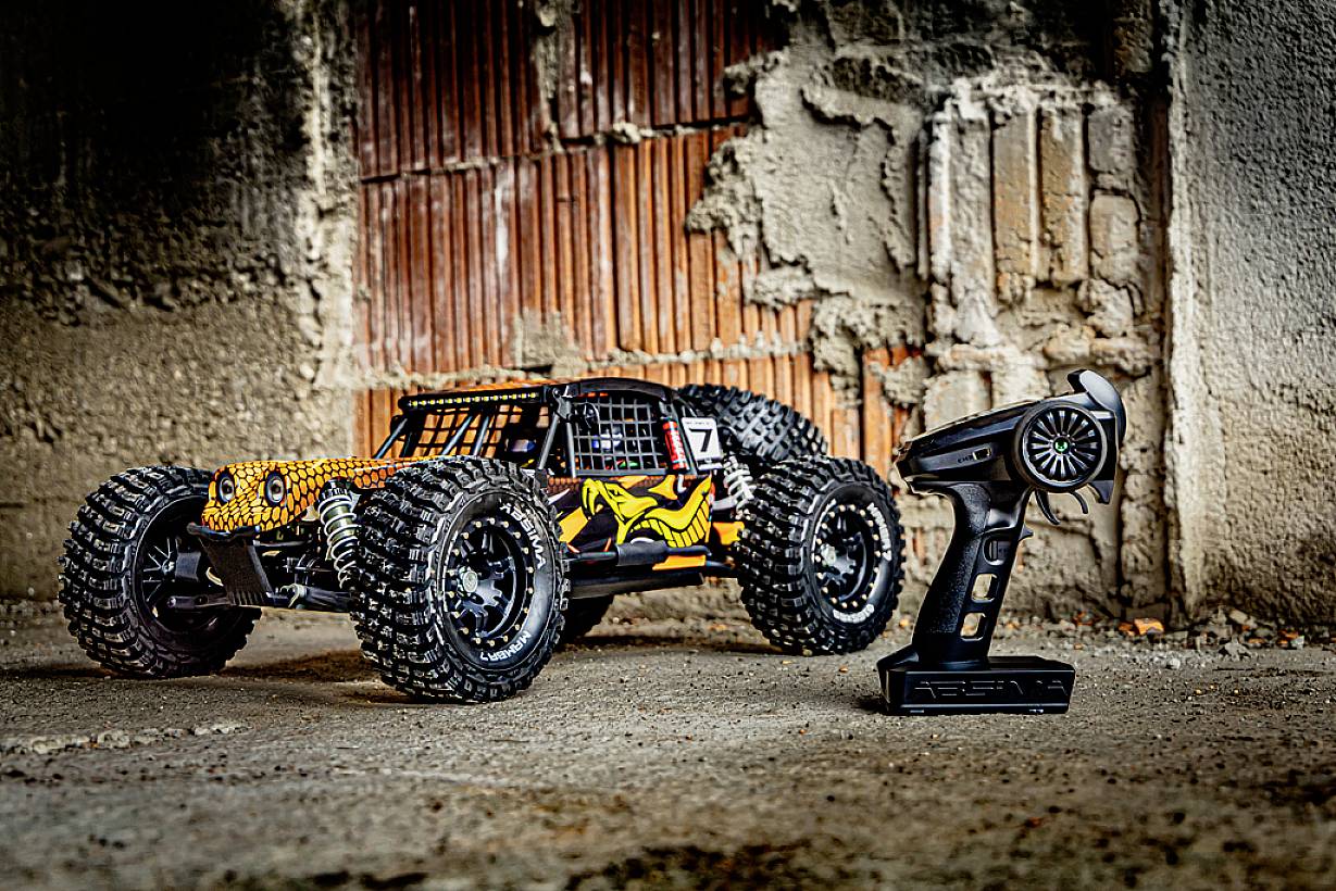 Ein ferngesteuertes Auto mit großen Reifen steht auf einem Betonboden neben einer Fernbedienung, vor einer rustikalen Ziegelmauer.