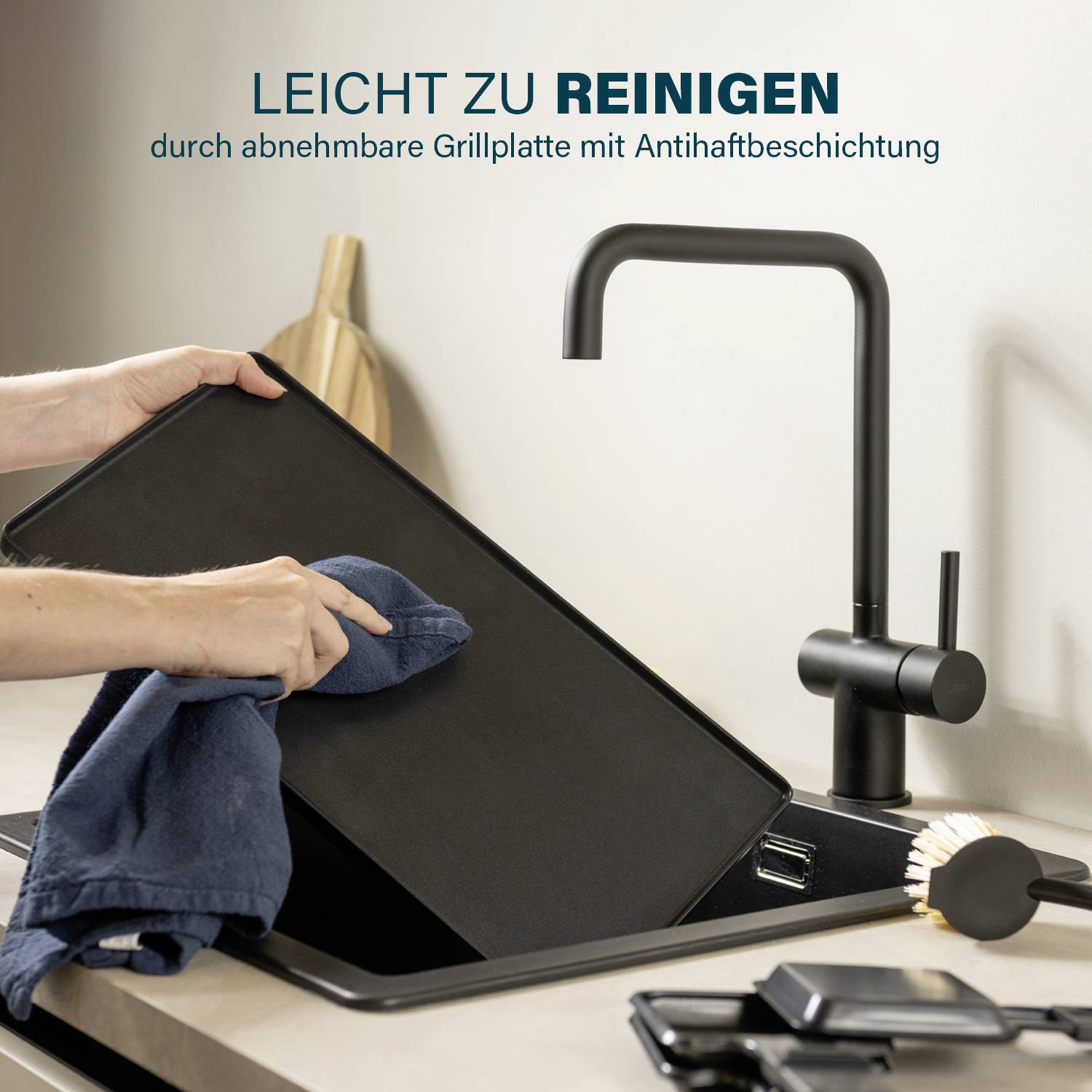 Person reinigt eine abnehmbare Grillplatte mit einem Tuch unter einem schwarzen Wasserhahn. Text: 'LEICHT ZU REINIGEN'.