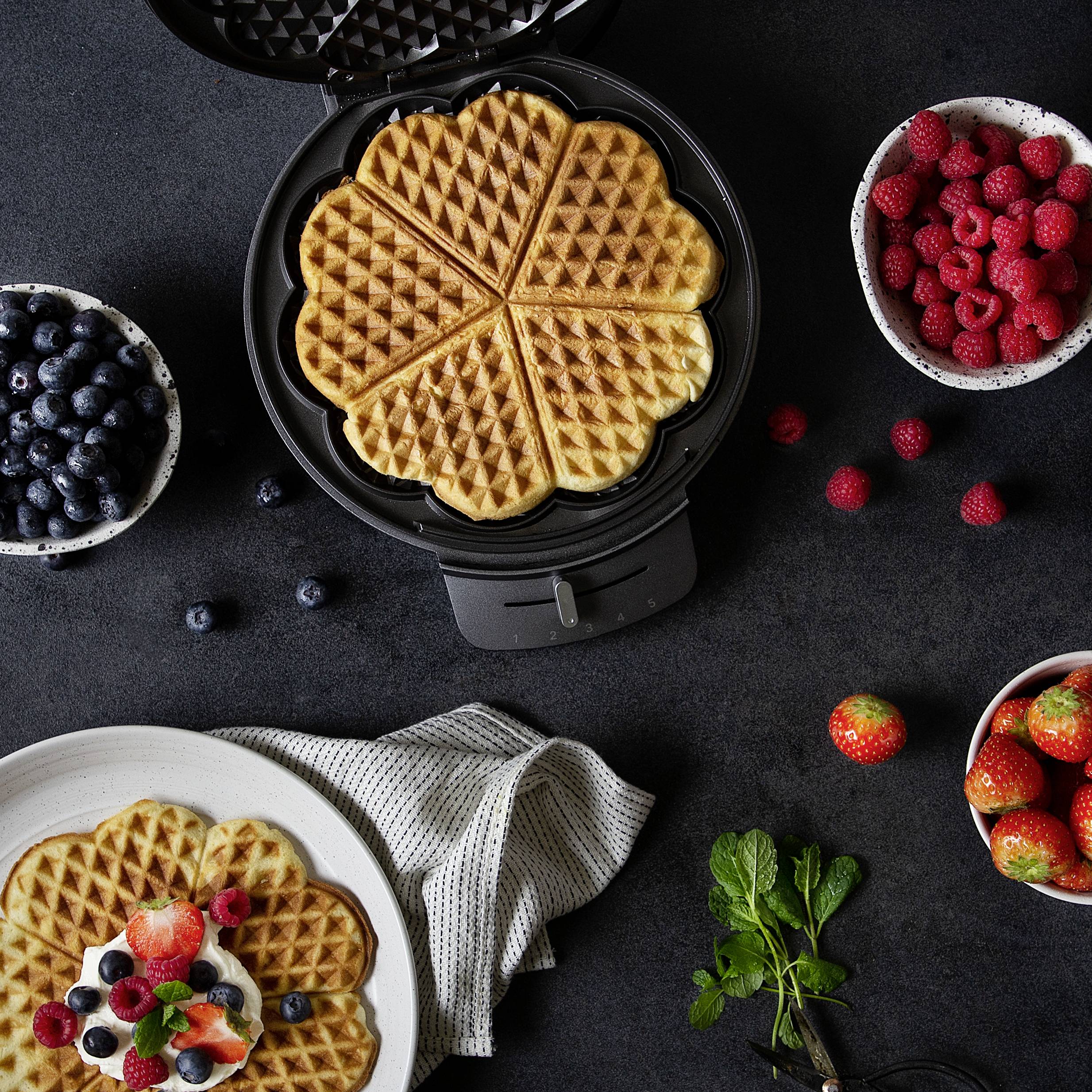 Ein Waffeleisen mit frischen Waffeln, umgeben von Schalen mit Blaubeeren, Himbeeren und Erdbeeren auf einem dunklen Tisch.