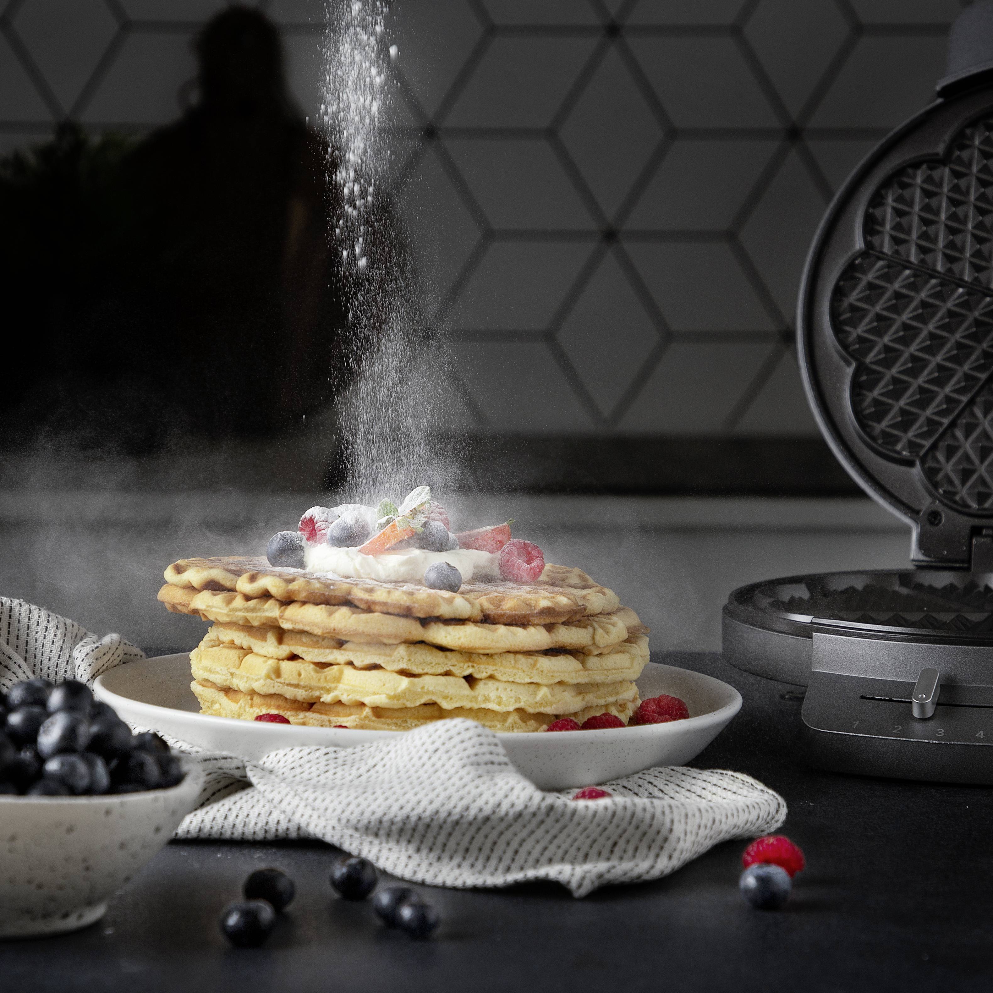 Ein Stapel Pfannkuchen mit Puderzucker, Himbeeren und Blaubeeren auf einem Teller. Ein Waffeleisen steht daneben auf der Arbeitsplatte.