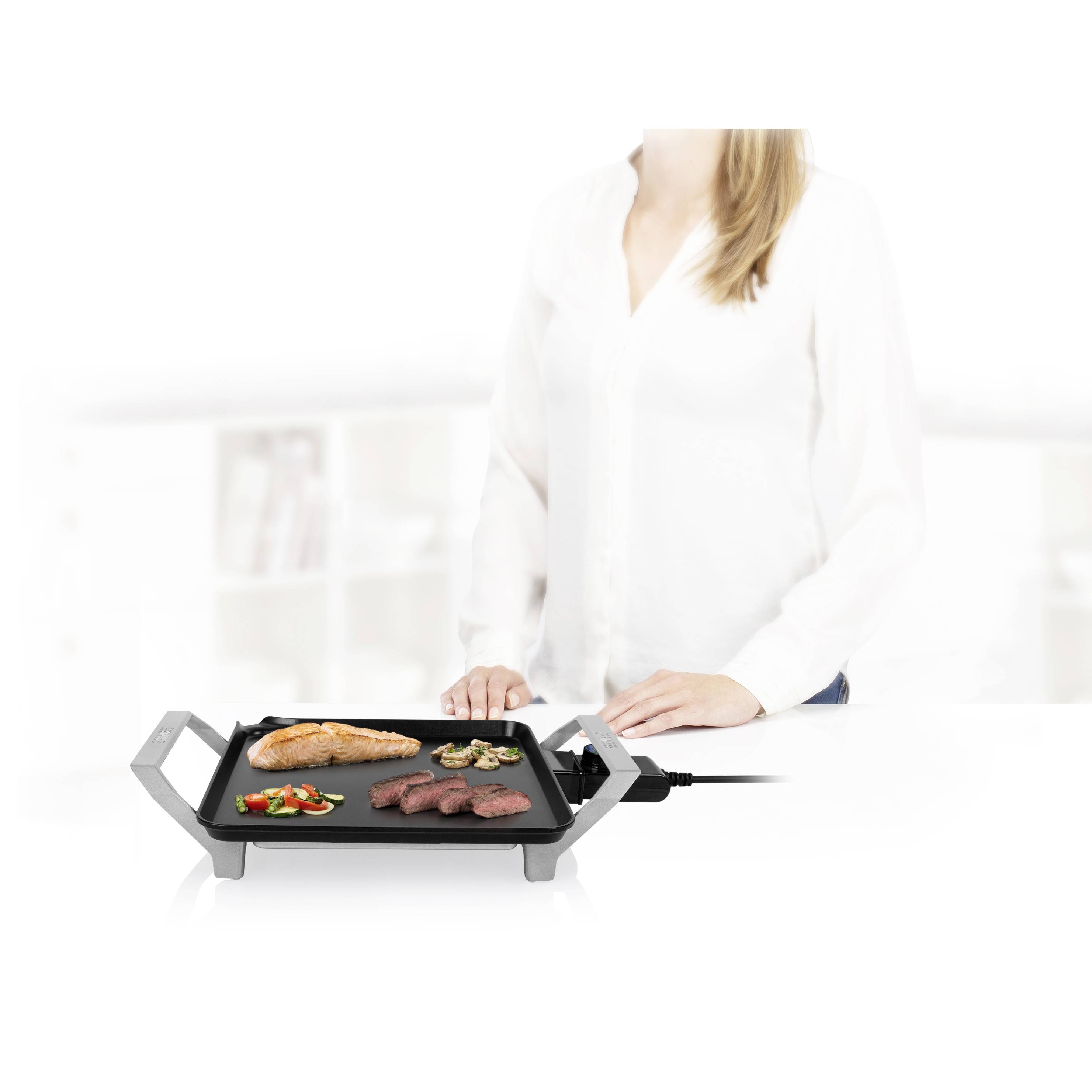 Eine Frau steht neben einem Tischgrill mit Fleisch, Fisch und Gemüse darauf, in einer modernen Küche.