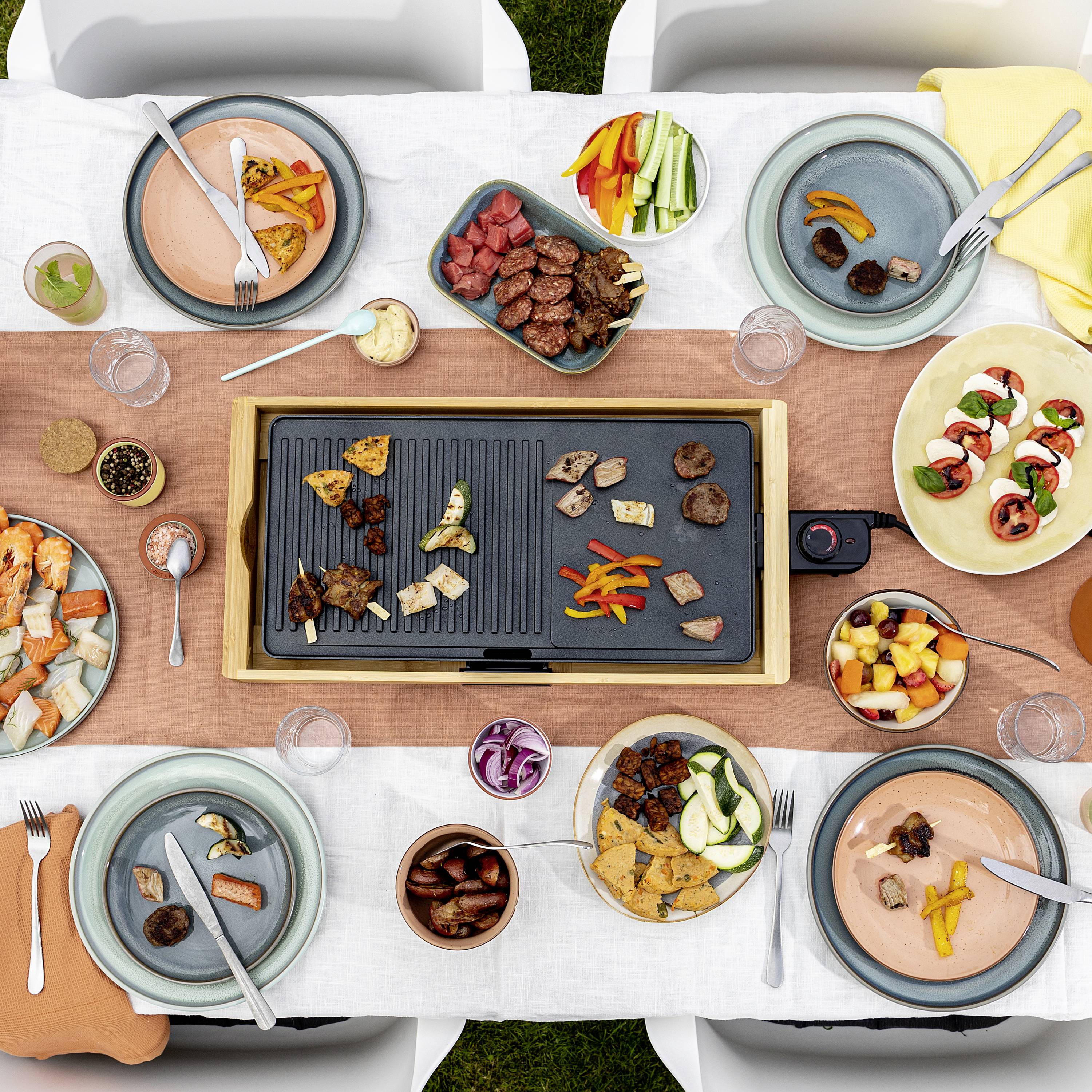 Eine Tischszene mit einer elektrischen Grillplatte in der Mitte, umgeben von verschiedenen Tellern mit Fleisch, Gemüse und Obst.