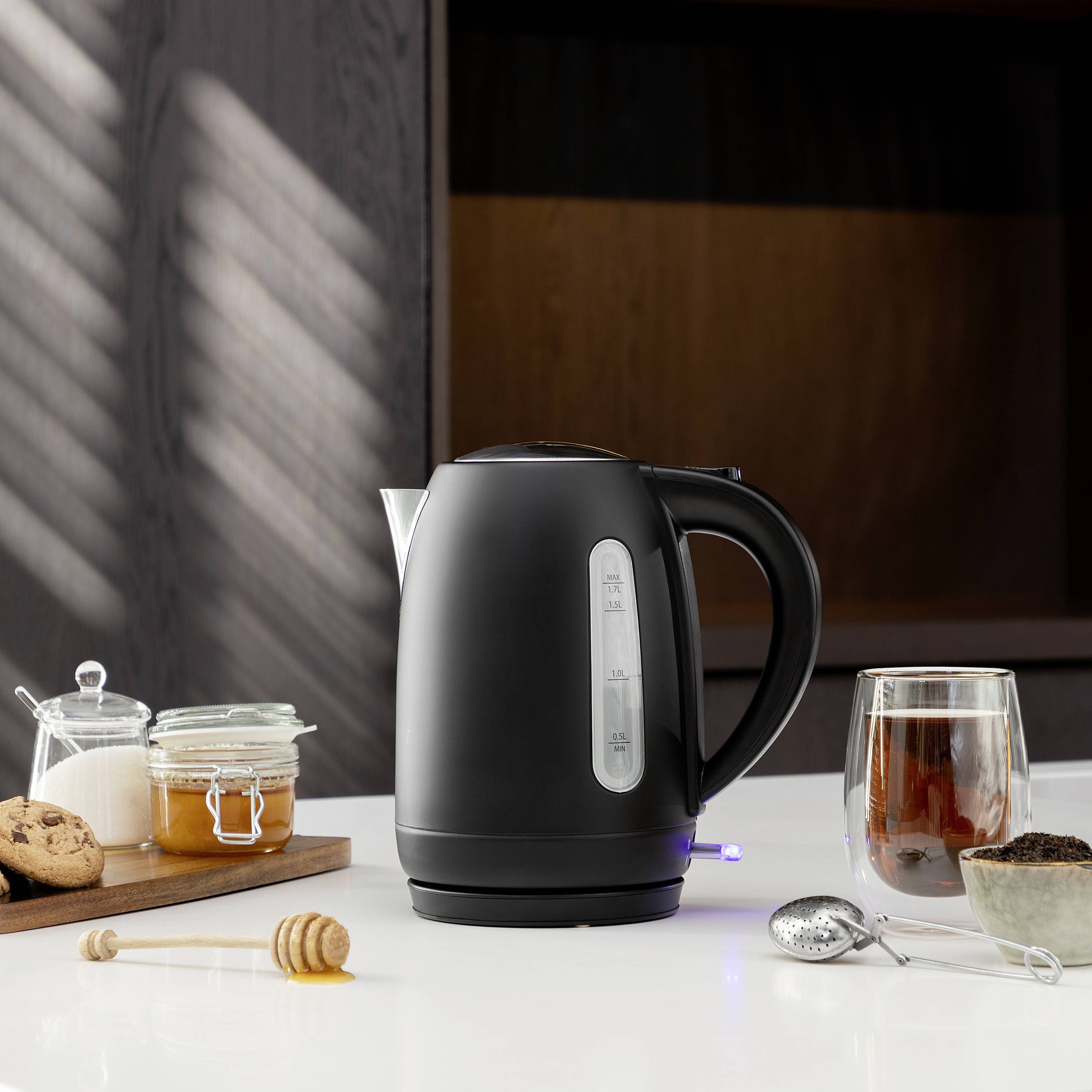 Ein schwarzer Wasserkocher steht auf einer Küchentheke neben einer Tasse Tee, einem Glas Honig und Keksen.
