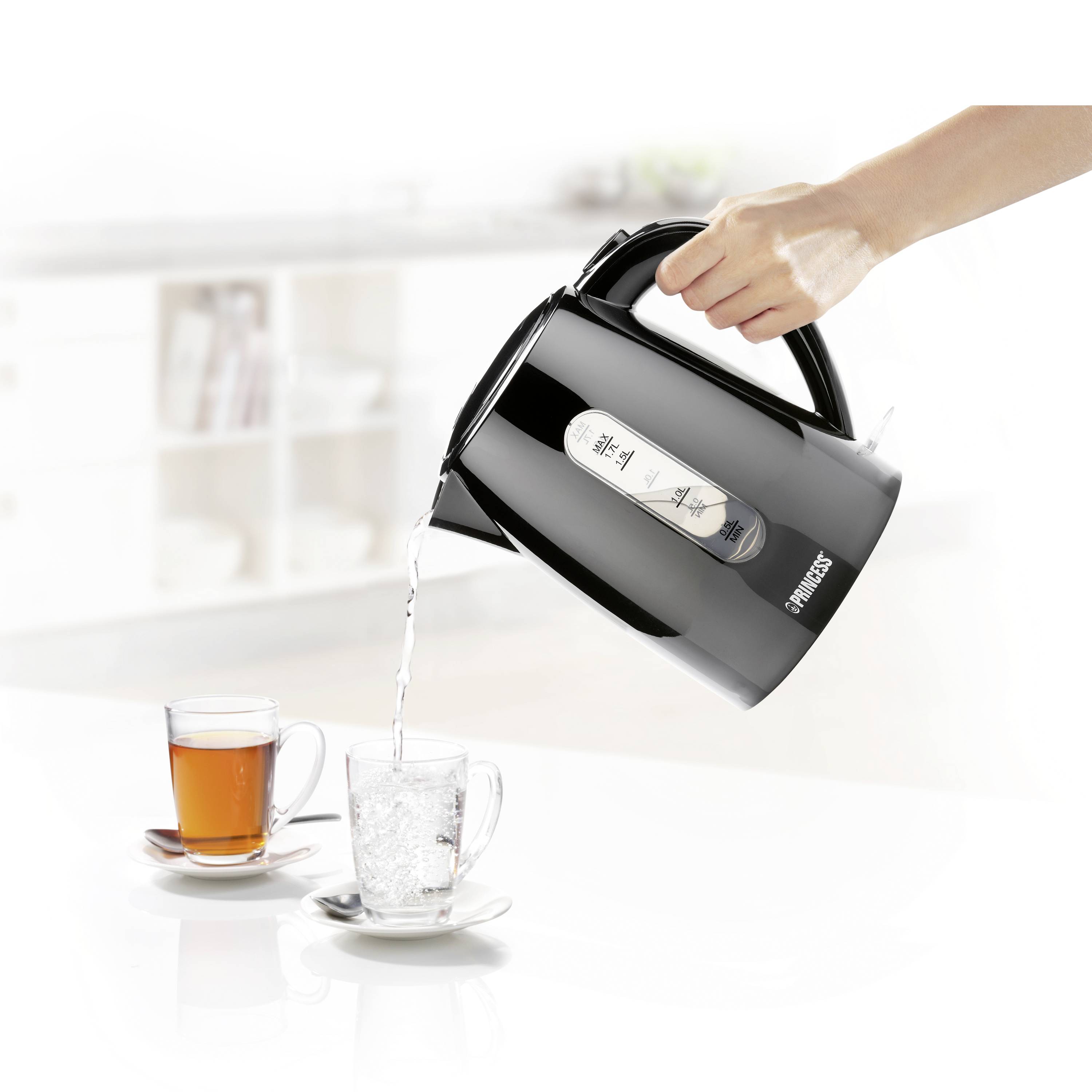 Eine Hand hält einen schwarzen Wasserkocher, der Wasser in eine Glastasse gießt. Im Hintergrund steht eine weitere Tasse mit Tee.