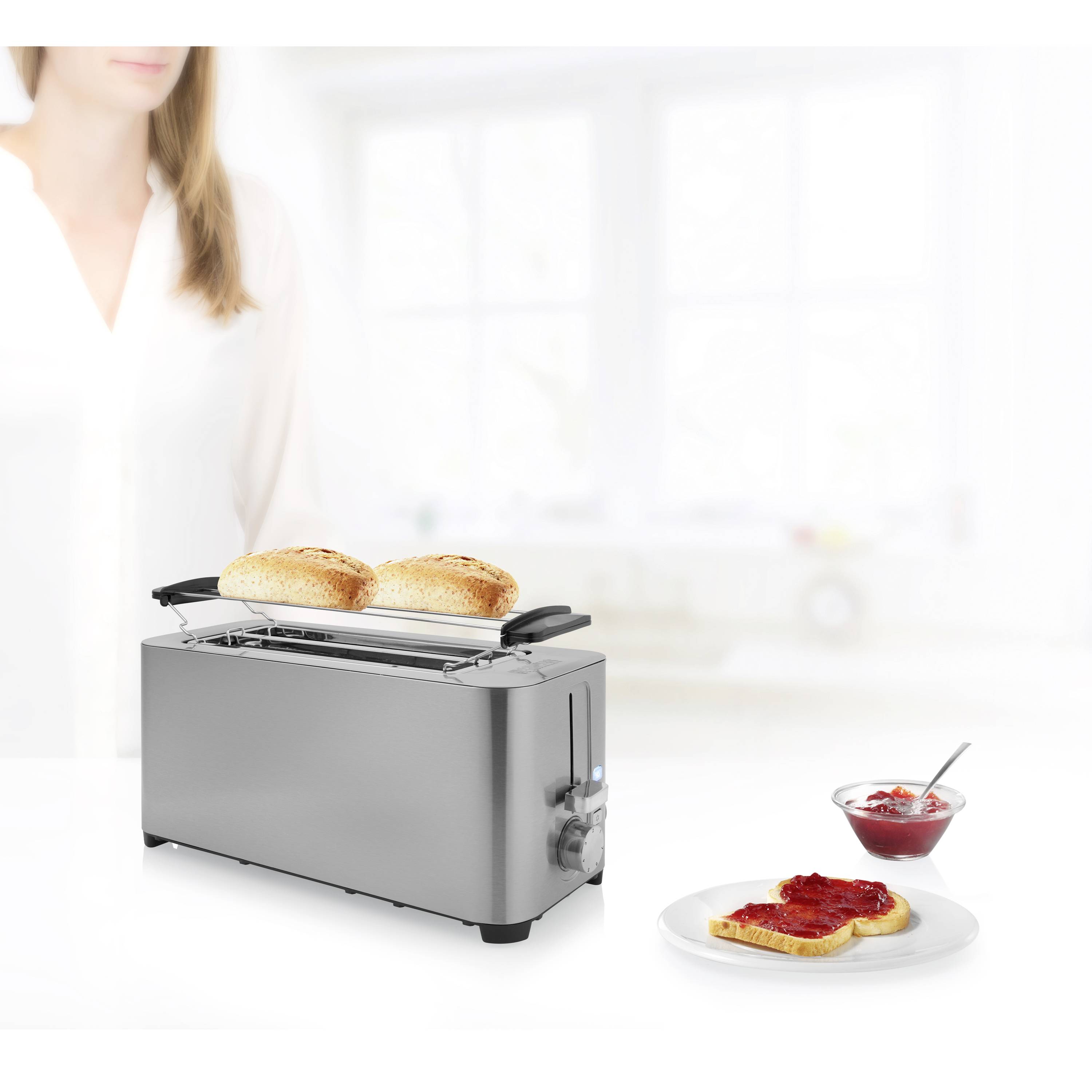 Ein silberner Toaster mit zwei Scheiben Toast neben einem Teller mit Toast und roter Marmelade. Im Hintergrund eine unscharfe Person.