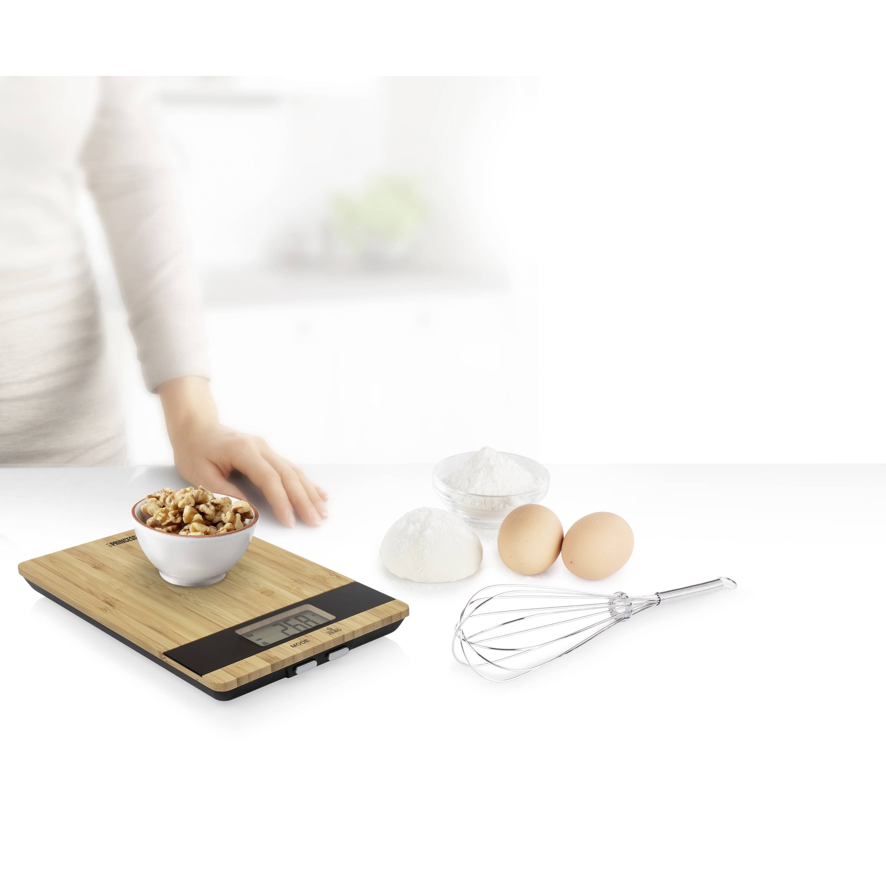 Frau wiegt Zutaten auf einer digitalen Küchenwaage: Schüssel mit Nüssen. Im Hintergrund Eier, Mehl in Schalen, und Schneebesen.