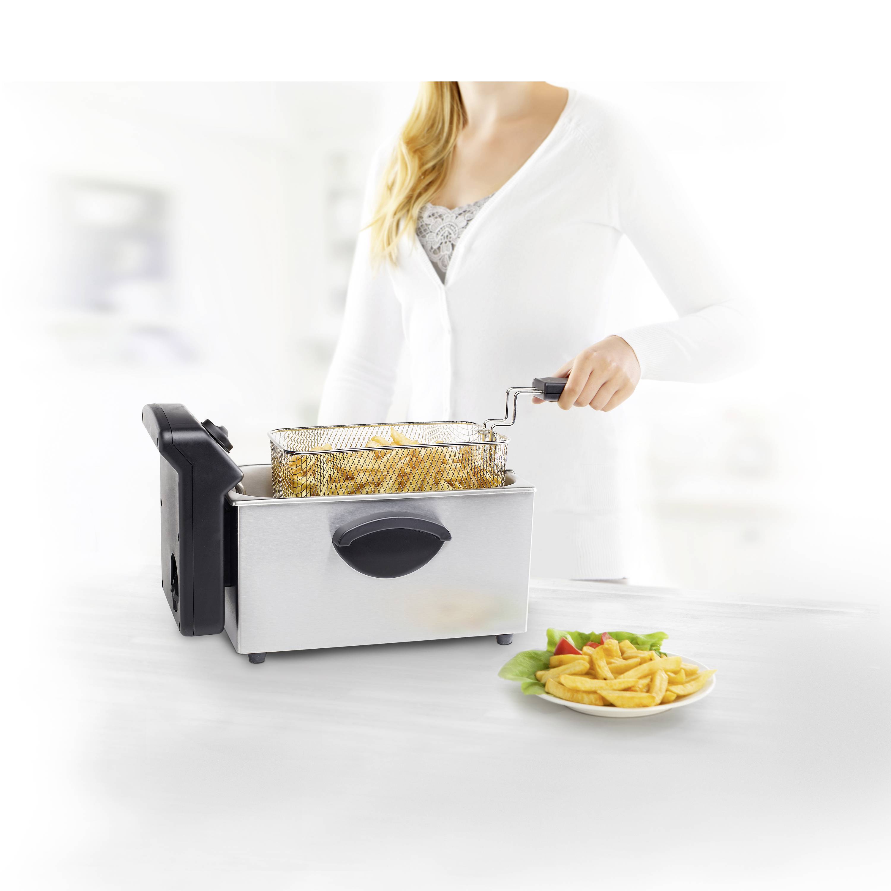 Eine Person hebt einen Korb mit frittierten Pommes aus einer modernen Edelstahl-Fritteuse. Daneben liegt ein Teller Pommes.