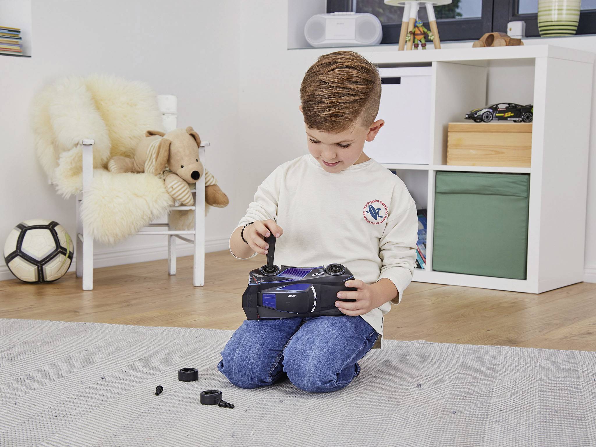 Ein Junge sitzt auf dem Boden und spielt mit einem ferngesteuerten Auto. Um ihn herum liegen Autoteile, und im Hintergrund sind Spielsachen zu sehen.