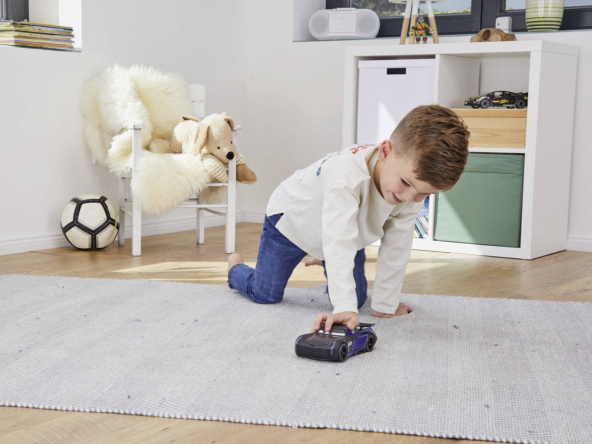 Ein Junge spielt fröhlich mit einem Spielzeugauto auf einem Teppich in einem hellen Kinderzimmer. Im Hintergrund ein Regal mit Spielzeug.