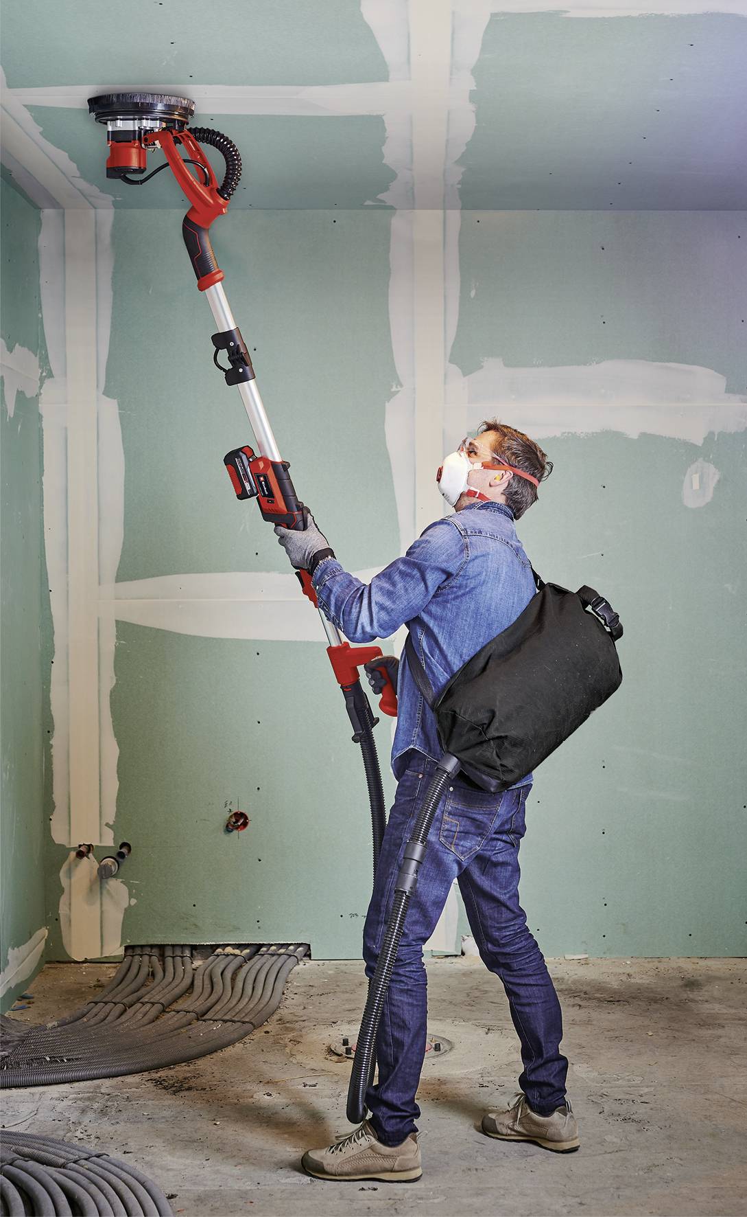 Person schleift Wand mit einer Schleifmaschine in einem renovierten Raum. Trägt Schutzmaske und hat Staubsauger-Rucksack.