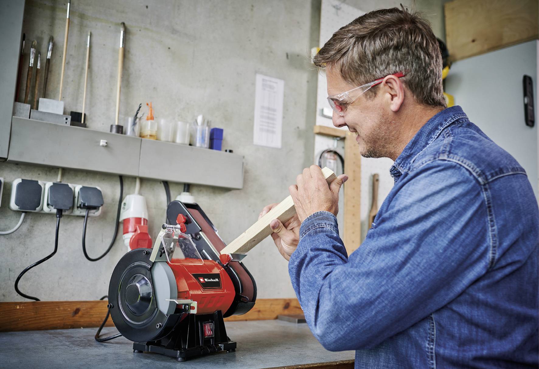 Ein Mann schleift ein Holzstück mit einem Bandschleifer in einer Werkstatt. Er trägt Schutzbrille und Jeanshemd.