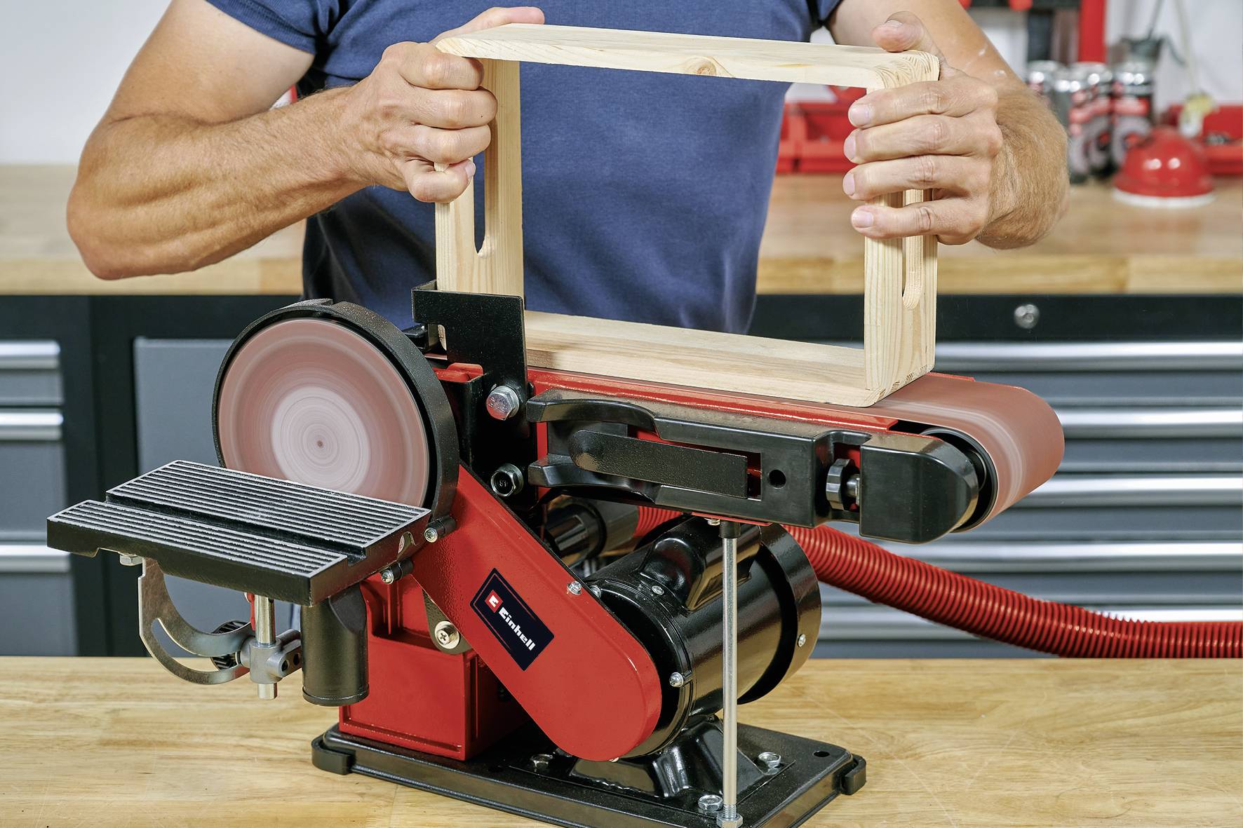 Person verwendet eine Bandschleifmaschine, um ein rechteckiges Holzstück zu bearbeiten. Schleifband und Arbeitstisch sichtbar.