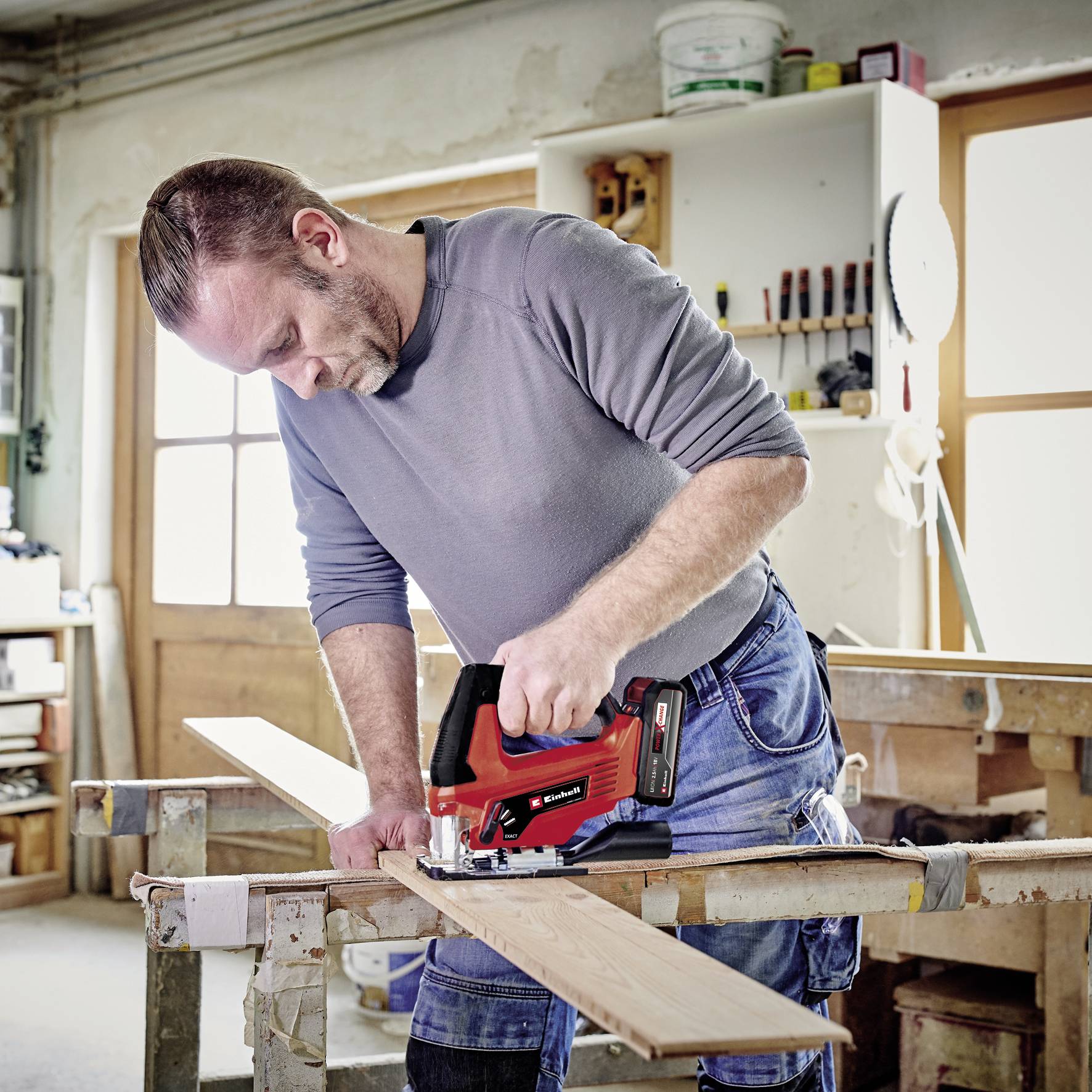 Ein Mann in einer Werkstatt bearbeitet ein Holzbrett mit einer elektrischen Säge. Im Hintergrund sind Werkzeugregale sichtbar.