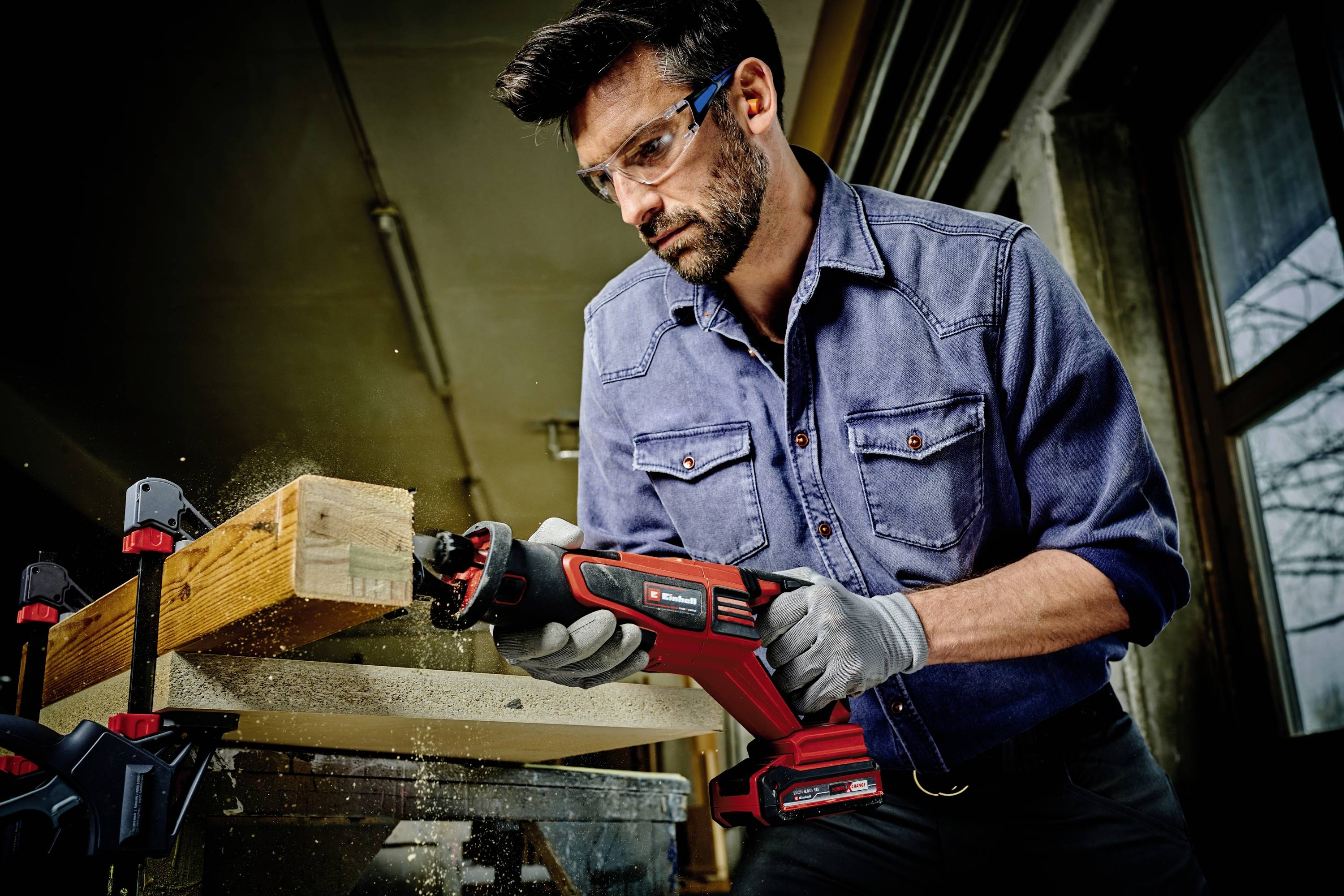Mann mit Schutzbrille und Handschuhen schneidet Holz mit einer elektrischen Säge in einer Werkstatt.