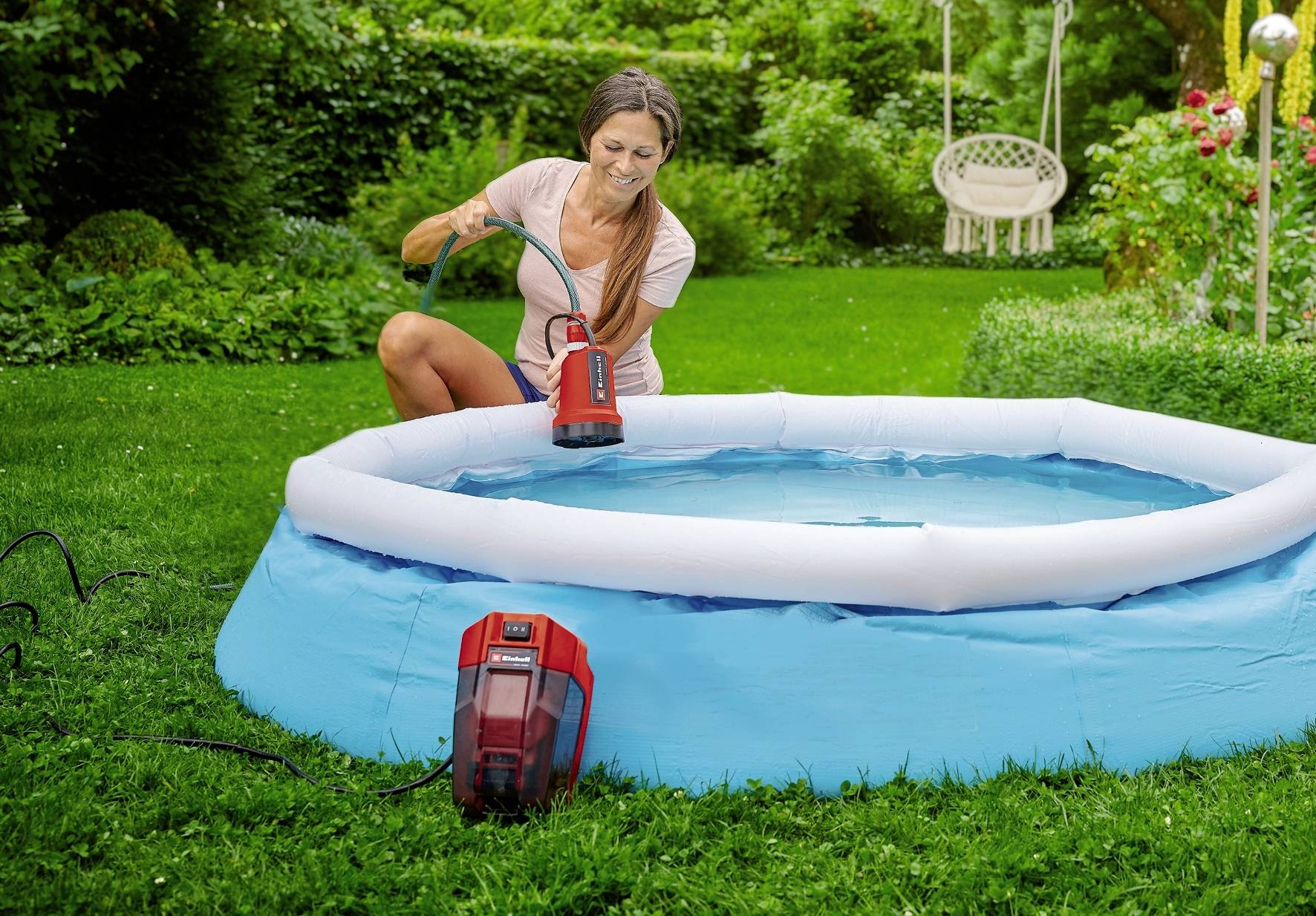 Eine Frau pumpt mit einem elektrischen Pumpgerät Wasser in einen aufblasbaren Pool im Garten. Im Hintergrund ist viel Grün zu sehen.