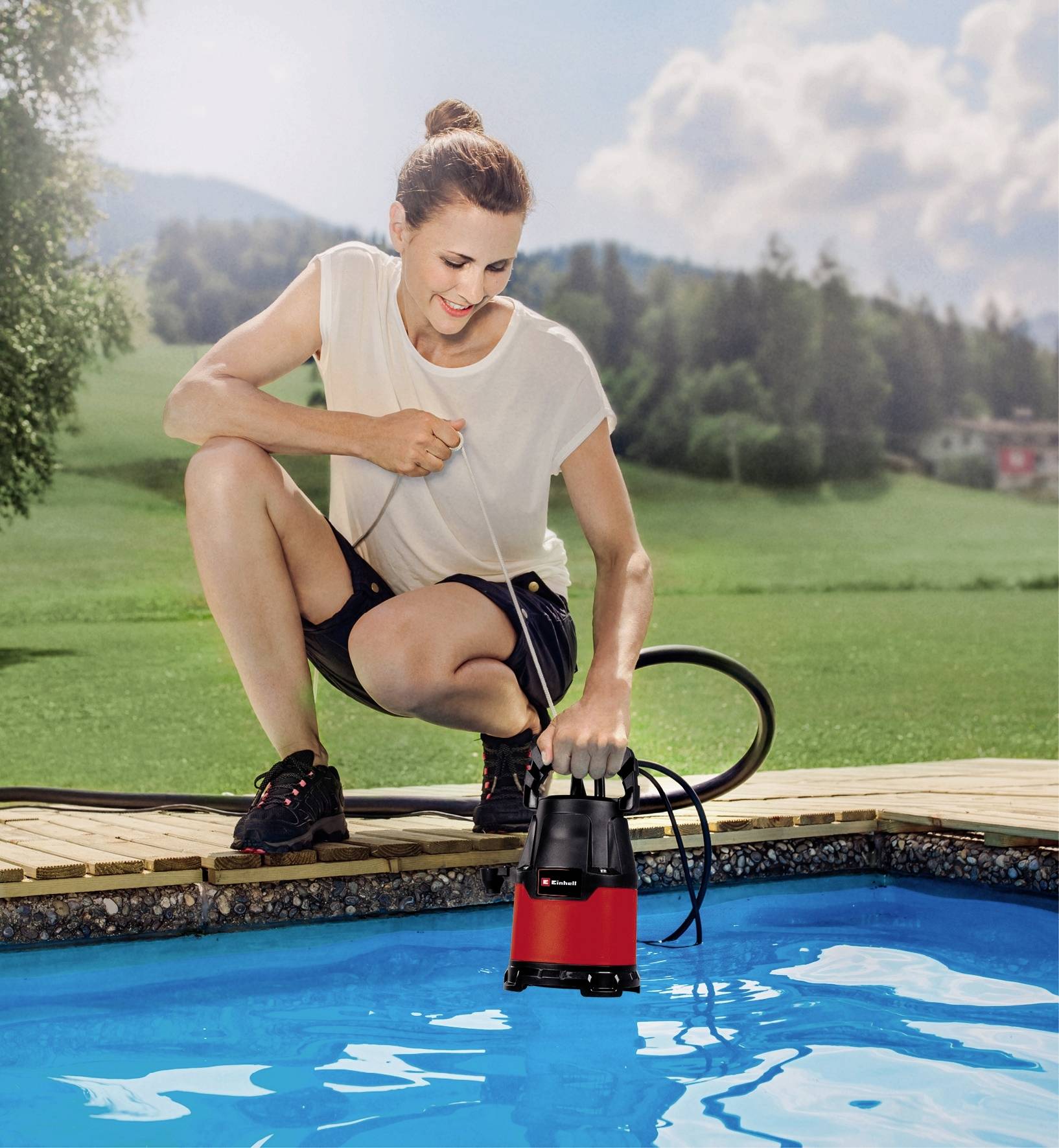 Eine Person kniet am Rand eines Pools und bedient eine Tauchpumpe, um Wasser abzupumpen. Im Hintergrund ist eine grüne Wiese.