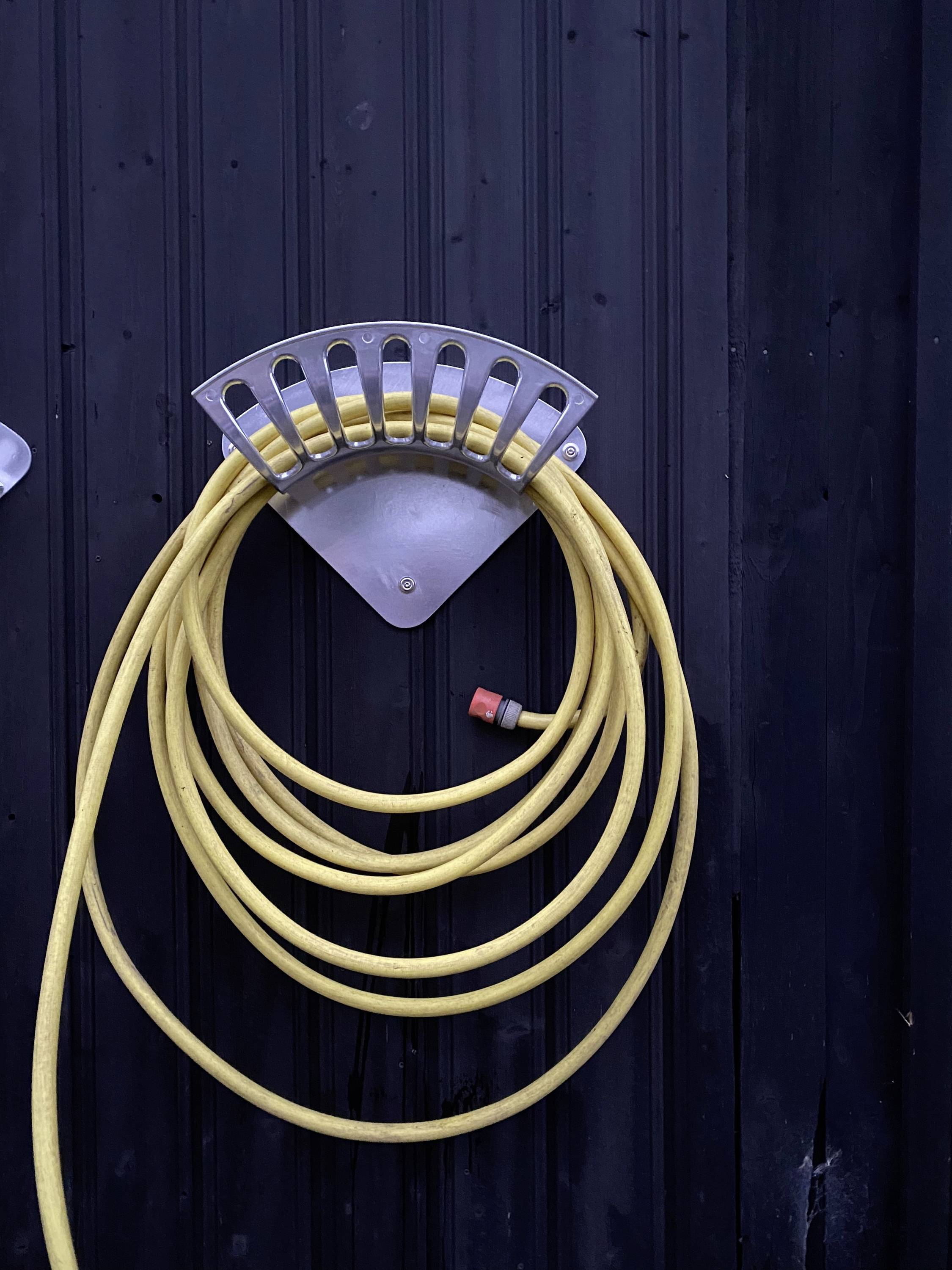 Ein gelber Gartenschlauch ist ordentlich an einem metallischen Wandhalter an einer schwarzen Wand aufgerollt.