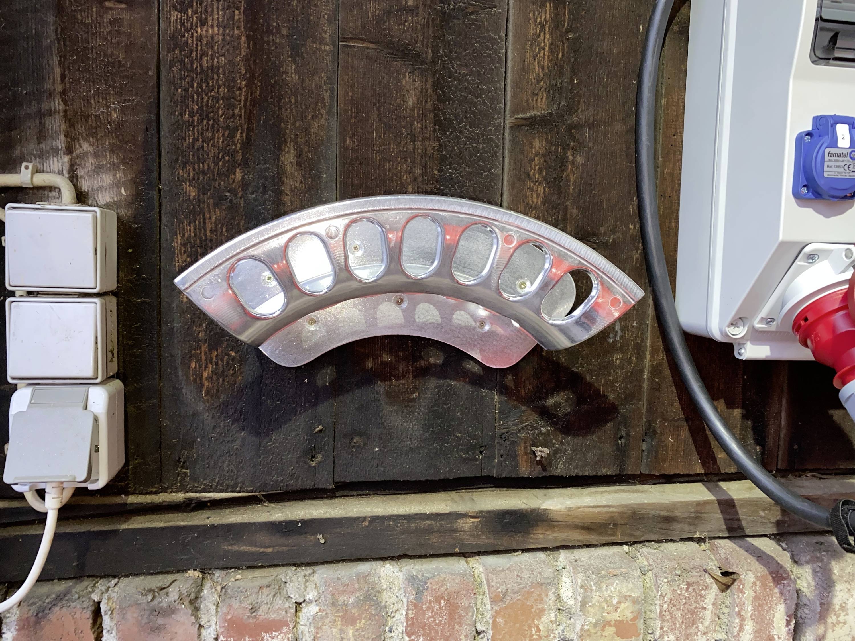 Metallhalterung an einer Holzwand montiert, umgeben von Steckdosen und Elektroinstallation, vor einer Backsteinmauer.