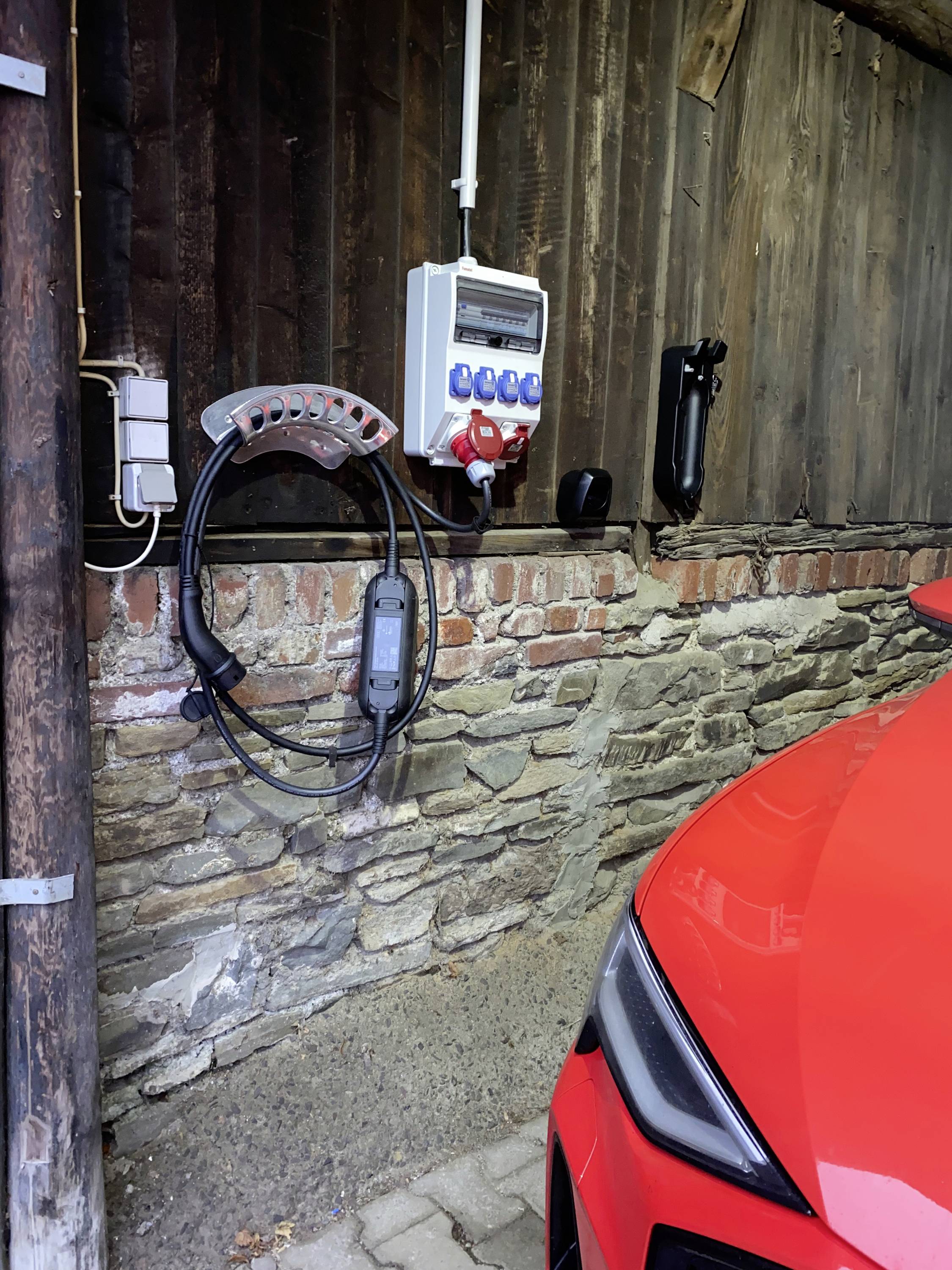 Rote Front eines Autos neben einer Wand mit einer Ladeeinrichtung. An der Wand sind eine Steckdose und ein Ladekabel befestigt.