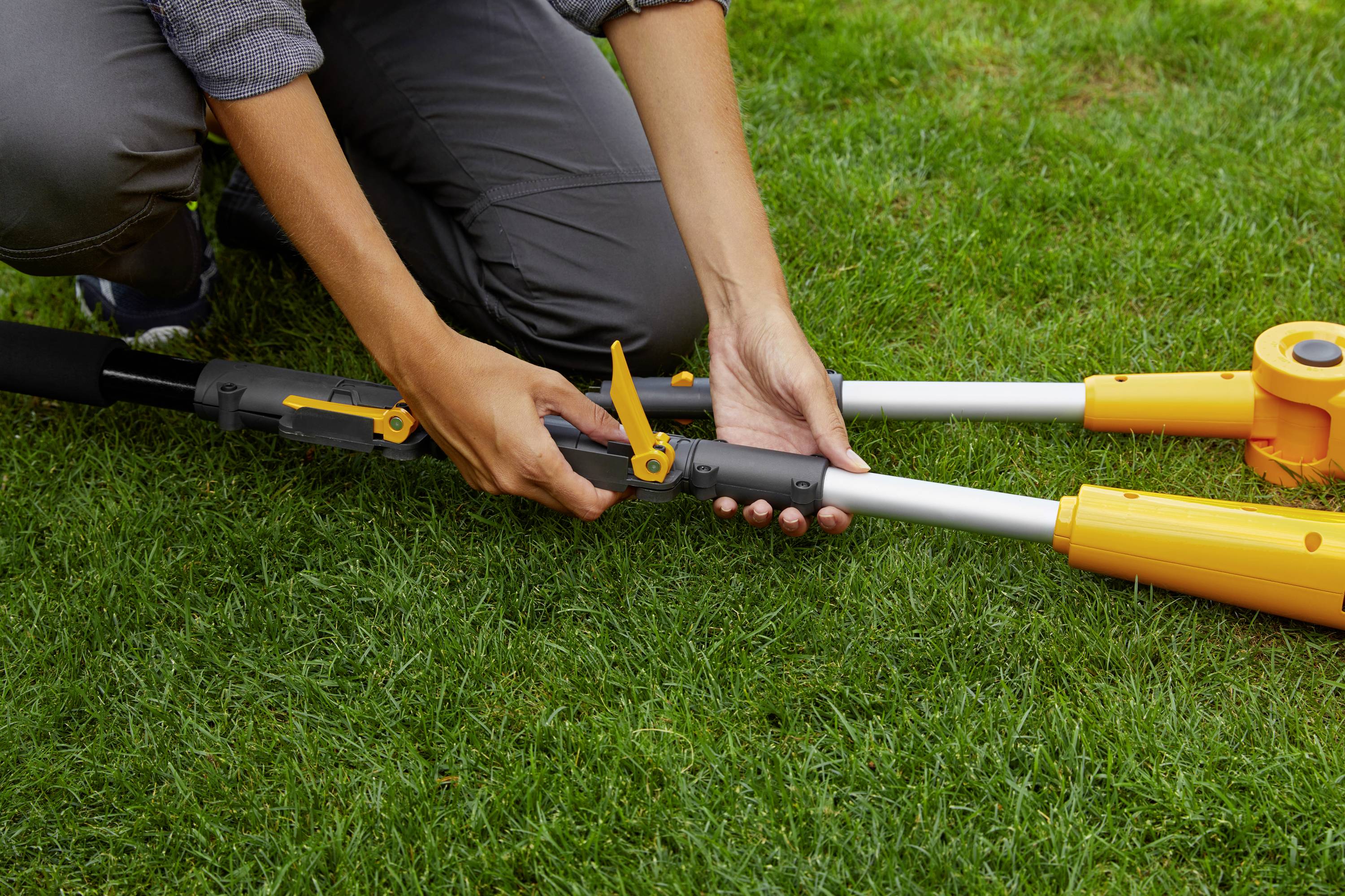 Eine Person montiert ein Gartengerät auf grünem Gras. Hände justieren einen Hebel an einem Gerät mit grauem und orangefarbenem Griff.