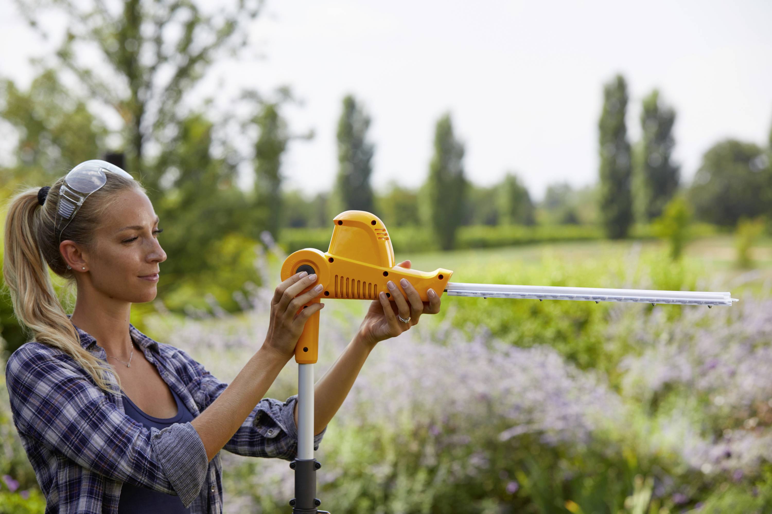 Eine Frau im Garten arbeitet mit einer elektrischen Heckenschere. Sie trägt Schutzbrille und ist von blühenden Pflanzen umgeben.