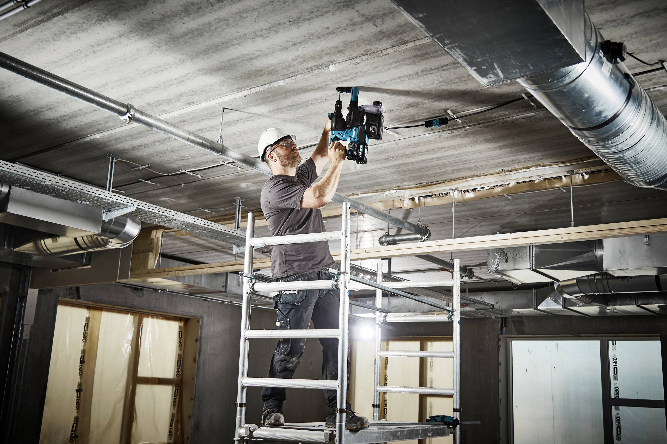 Ein Bauarbeiter steht auf einem Gerüst und bohrt in die Decke eines unfertigen Gebäudes. Er trägt einen Schutzhelm und benutzt eine elektrische Bohrmaschine.