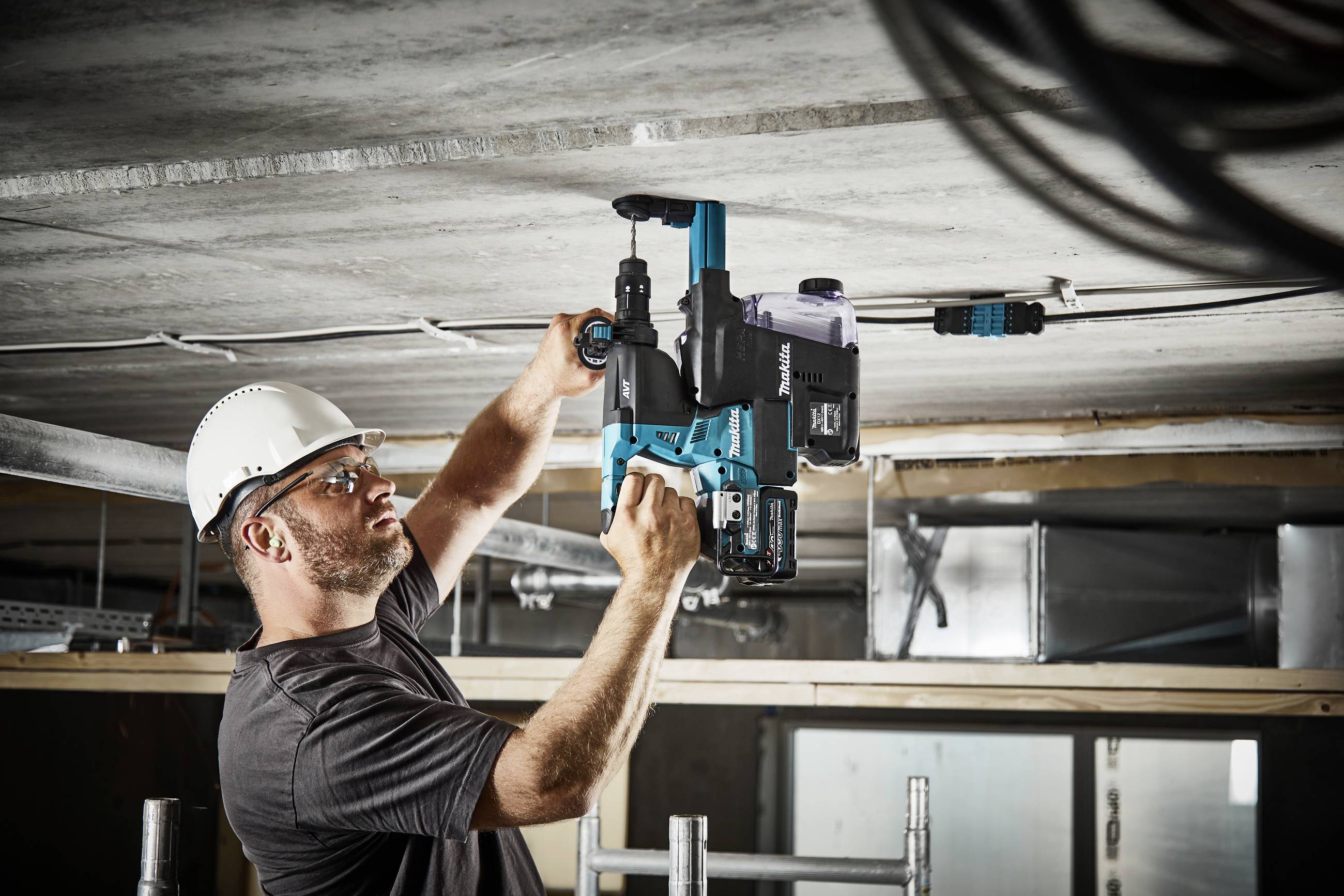 Ein Bauarbeiter mit Schutzhelm bohrt mit einer Elektrowerkzeug an einer Betondecke. Kabel und Baugerüste im Hintergrund.