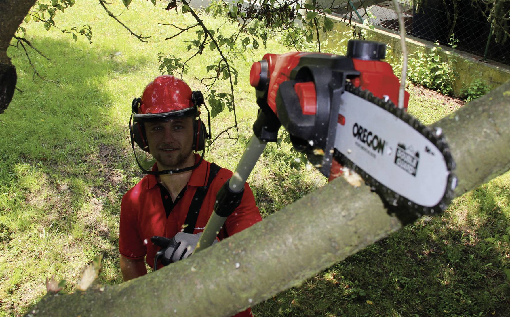 Ein Mann in rotem Helm und Shirt benutzt eine elektrische Kettensäge, um einen Ast eines Baumes in einem grünen Garten zu schneiden.