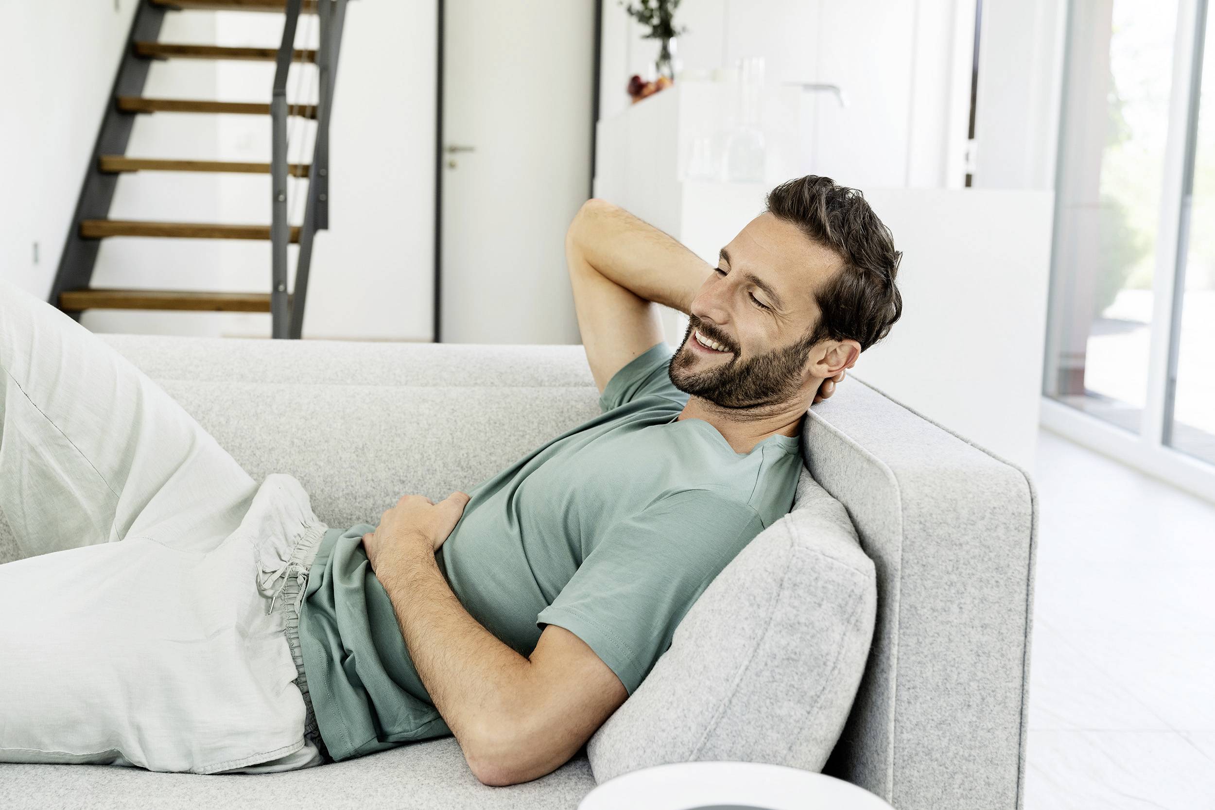 Ein Mann liegt entspannt auf einem grauen Sofa und lächelt, mit den Händen hinter dem Kopf verschränkt. Im Hintergrund ist eine Treppe zu sehen.