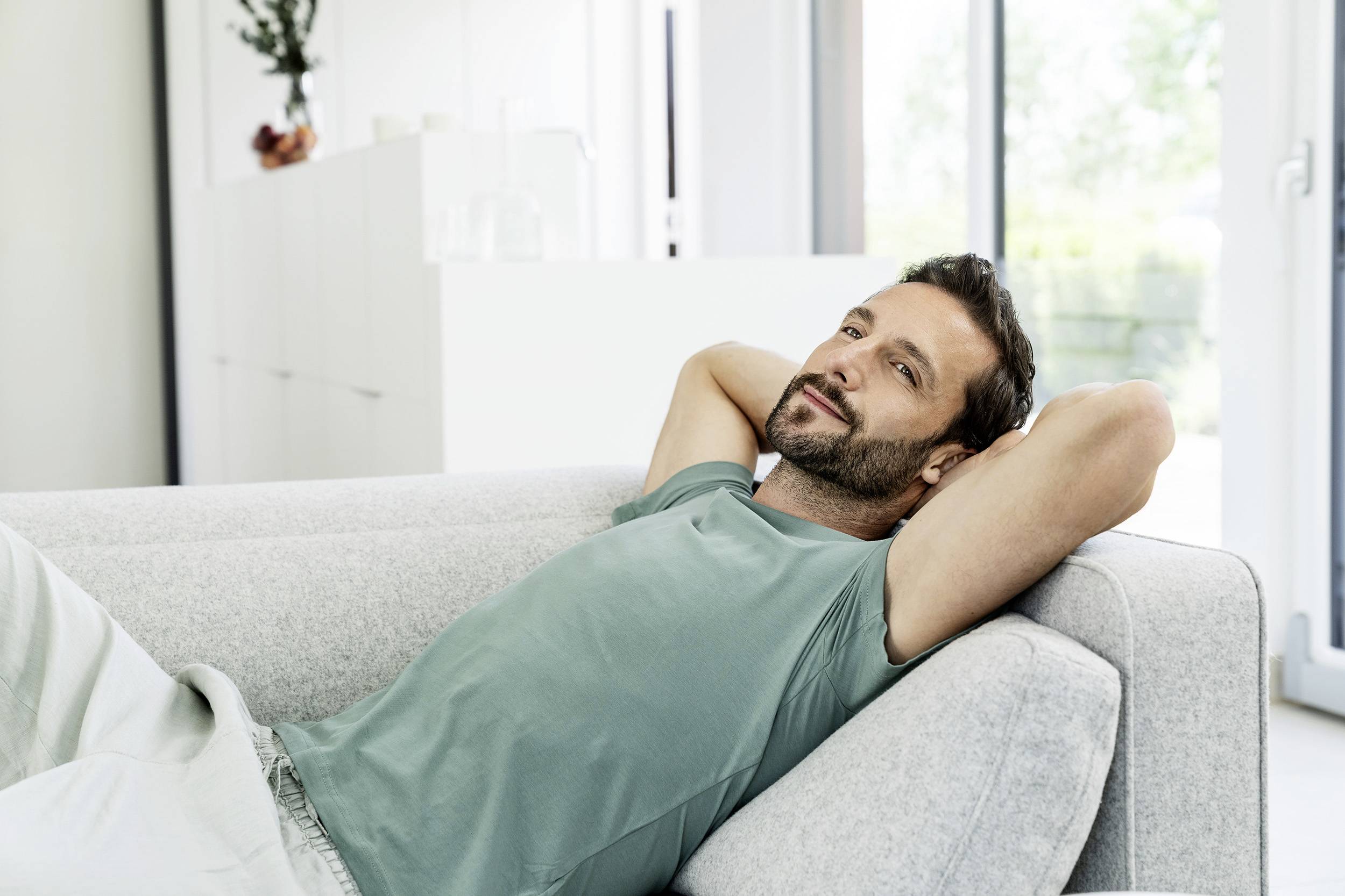 Ein Mann liegt entspannt auf einer Couch, mit den Händen hinter dem Kopf. Er trägt ein helles T-Shirt und blickt lächelnd zur Seite. Im Hintergrund sind helle Möbel und ein Fenster zu sehen.