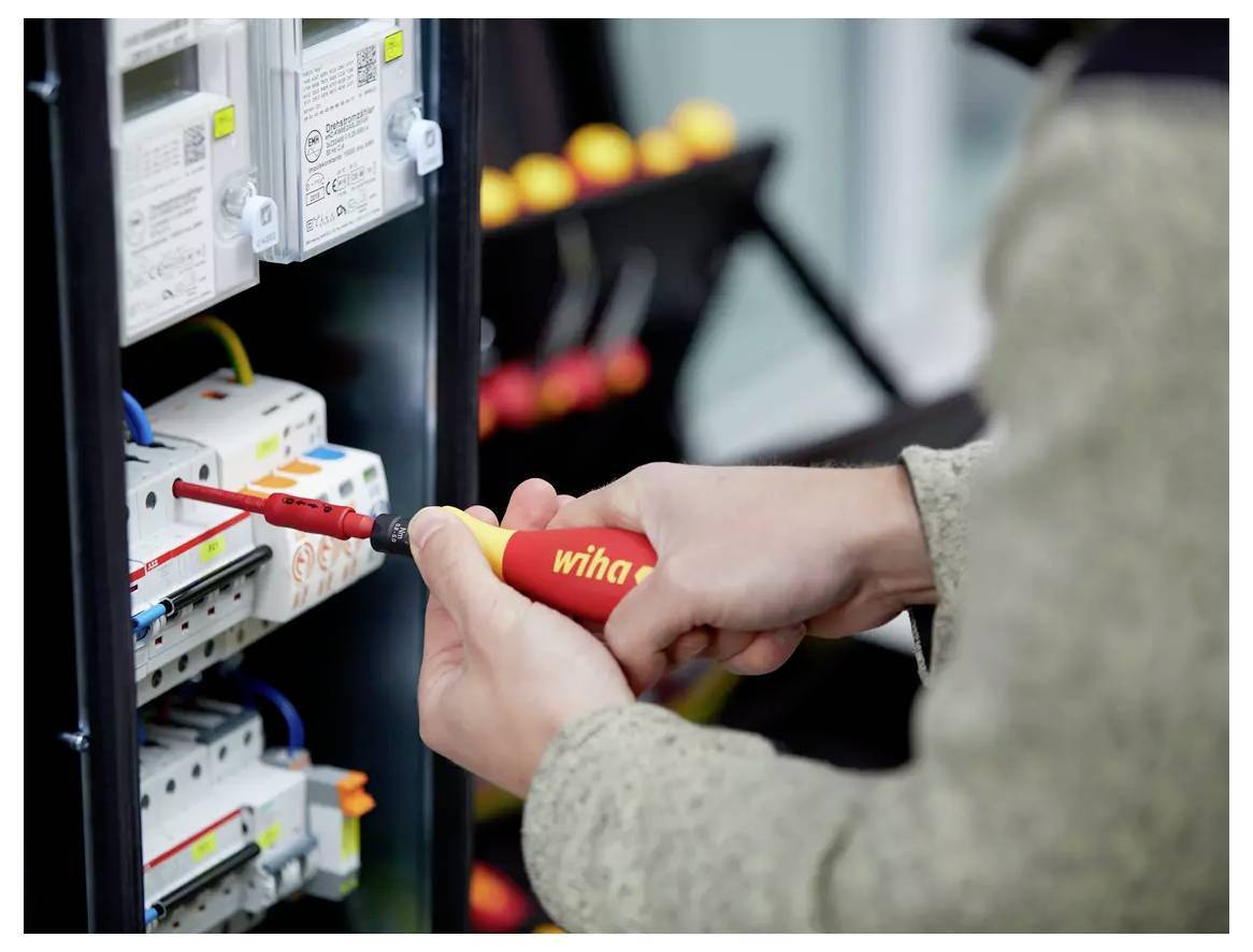 Eine Person verwendet einen rot-gelben Schraubendreher, um elektrische Verbindungen in einer Schutzschaltertafel anzupassen.