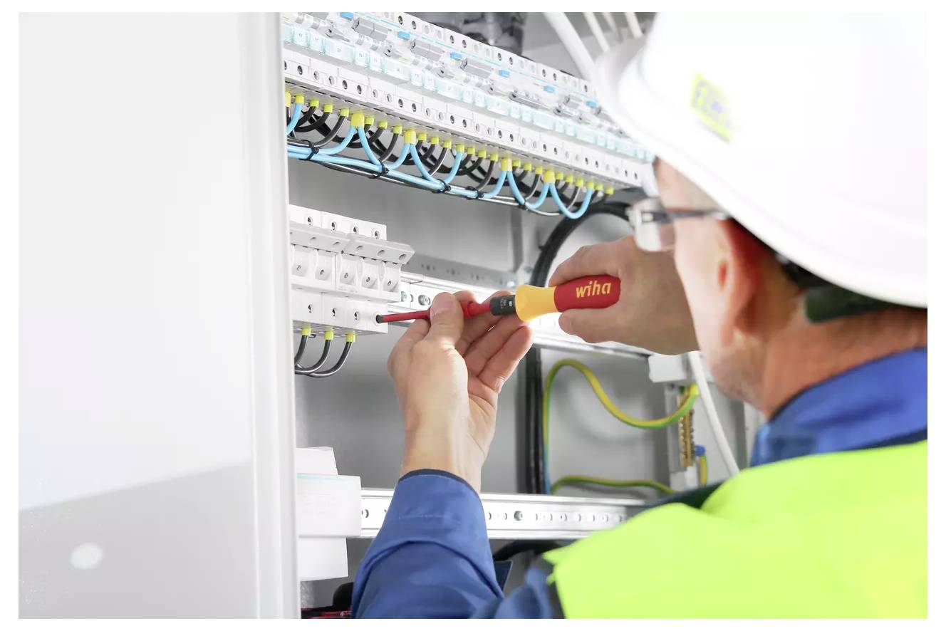 Ein Elektriker, der einen Schutzhelm und eine Sicherheitsweste trägt, benutzt einen Schraubendreher, um die Verdrahtung in einem Schaltschrank zu justieren.