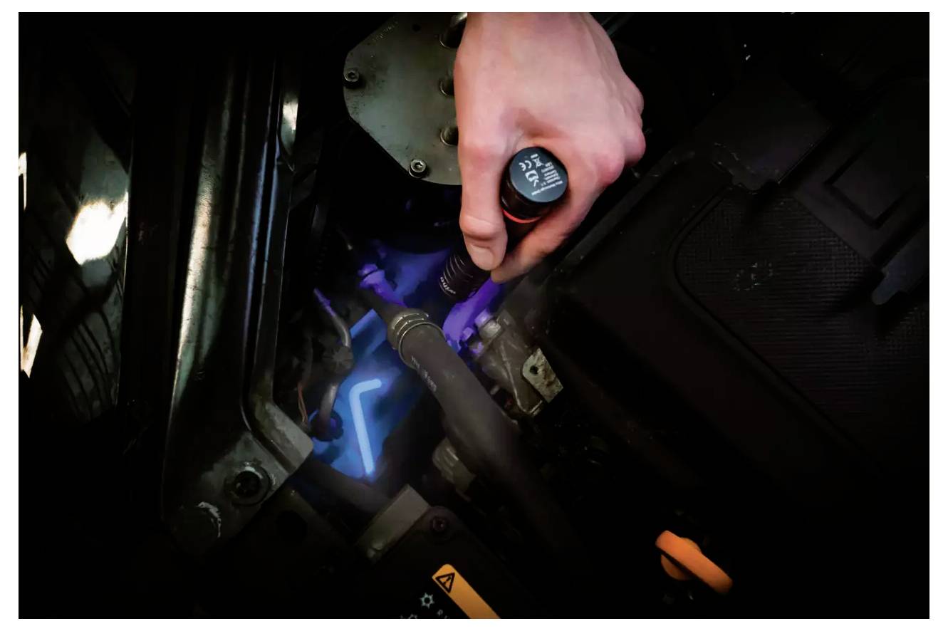 Hand, die einen Ölmessstab über einem Automotor hält, um den Ölstand zu überprüfen. Motorteile sind sichtbar, beleuchtet durch neonblaue Beleuchtung.