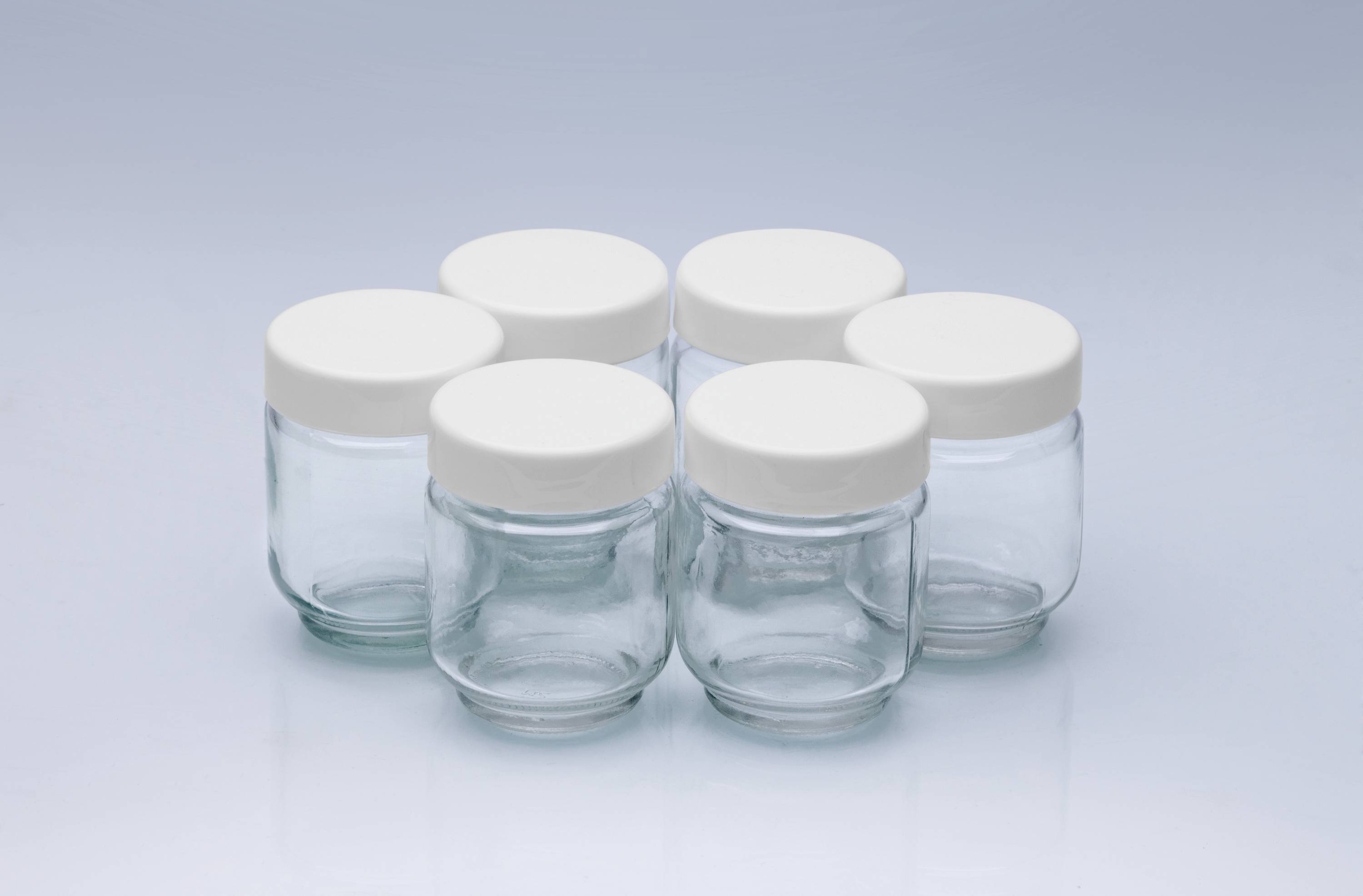 Sechs leere, gläserne Einmachgläser mit weißen Deckeln stehen auf einem hellblauen Hintergrund in einem Kreis angeordnet.