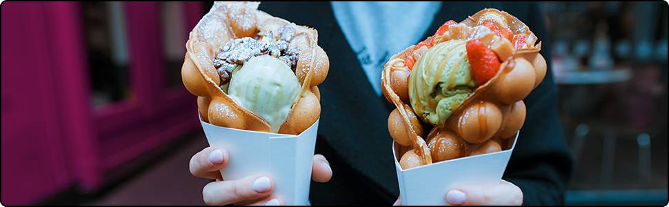 Zwei Hände halten Waffelkegel mit runden Blasen. Jede Waffel enthält Eiscreme und Früchte als Topping. Hintergrund unscharf.