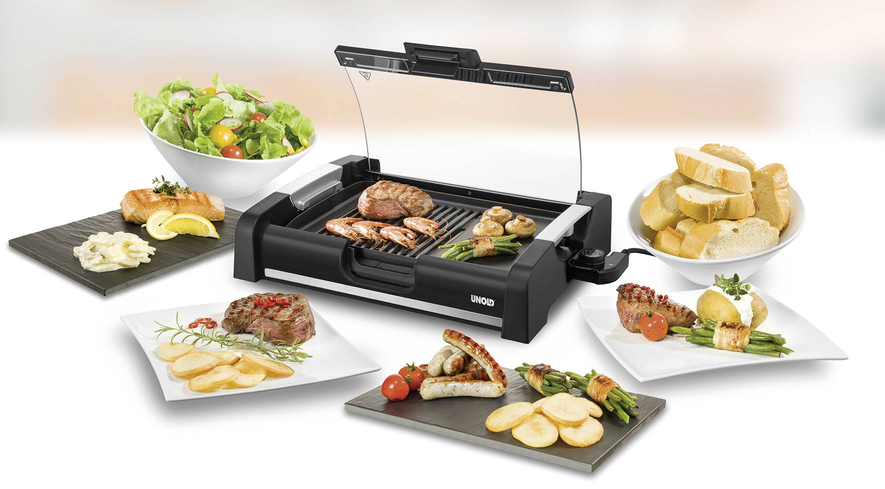 Ein Tischgrill mit gegrilltem Fleisch, Würstchen und Gemüse. Daneben Teller mit Brot, Salat, Kartoffeln und Garnelen.