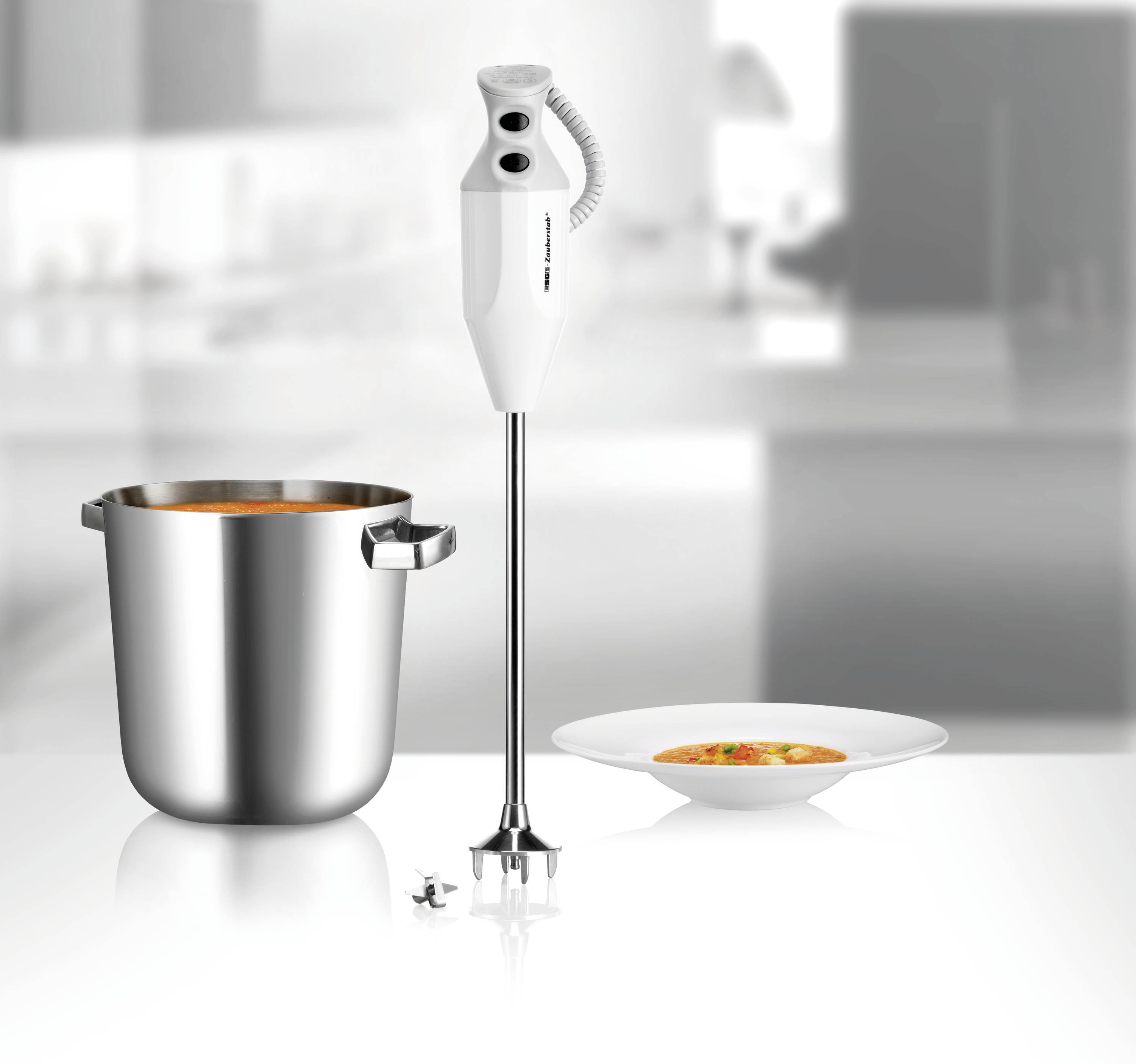 Pürierstab neben einem Topf mit Suppe und einem gefüllten Teller auf einem Tisch in einer modernen Küche.