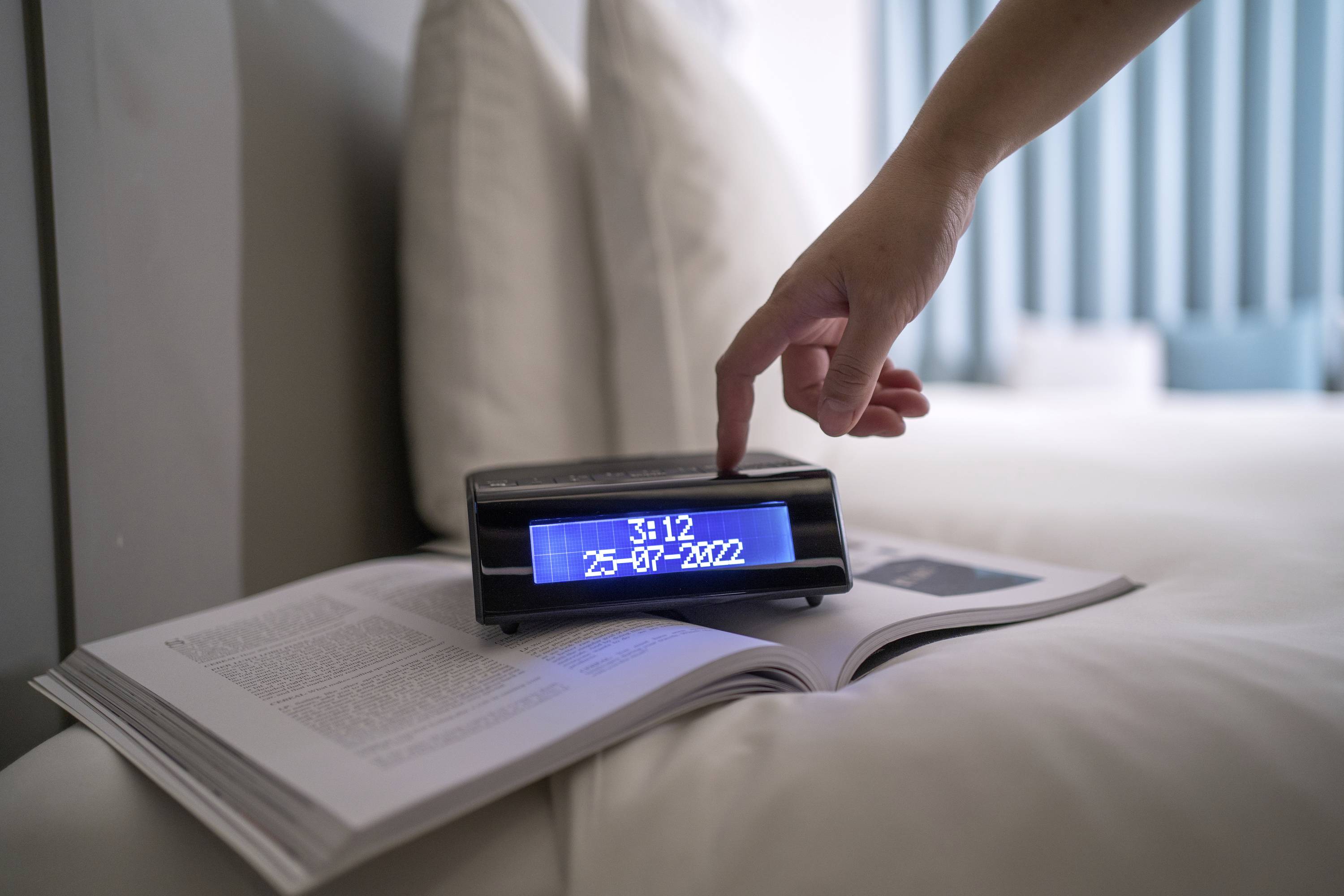 Eine Hand stellt einen digitalen Wecker auf einem geöffneten Buch ein. Der Wecker zeigt '25-07-2022' und eine Uhrzeit an.