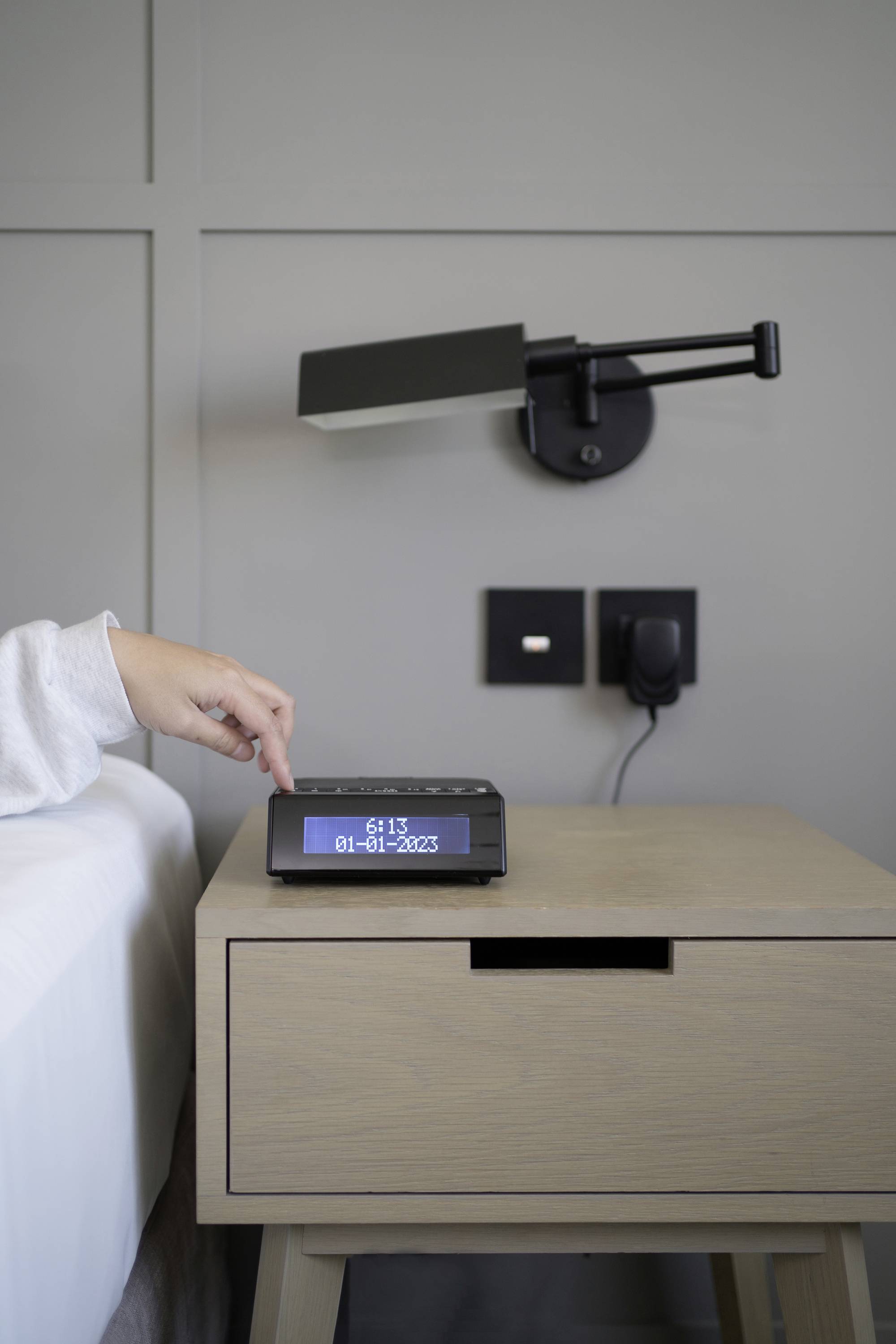 Eine Hand auf einem Nachttisch stellt einen Wecker ein. Der Tisch ist neben einem Bett, eine Lampe ist an der Wand montiert.